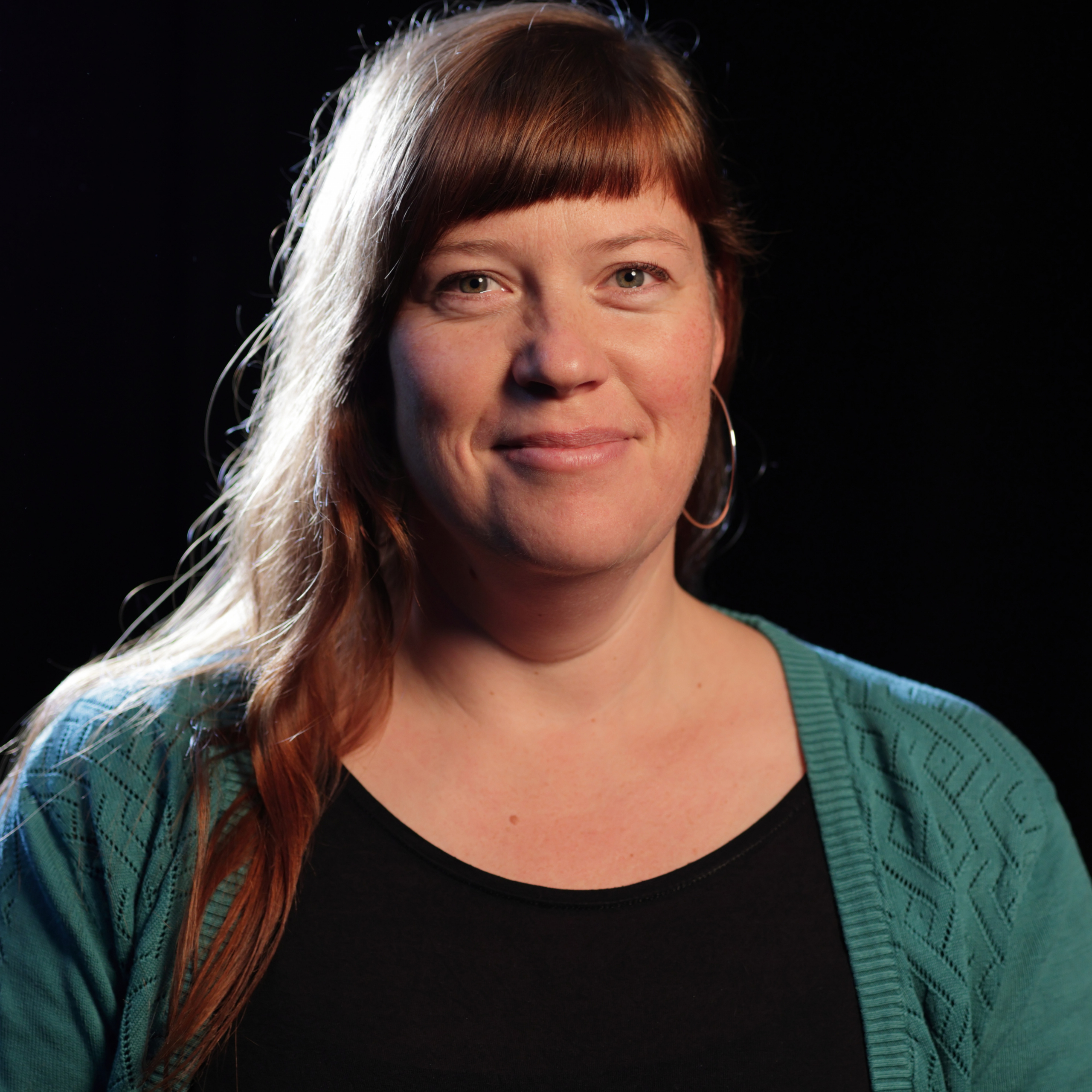 Frau mit langen, gewellten Haaren, schwarzem Oberteil und türkisfarbenem Strickjacke vor schwarzem Hintergrund.