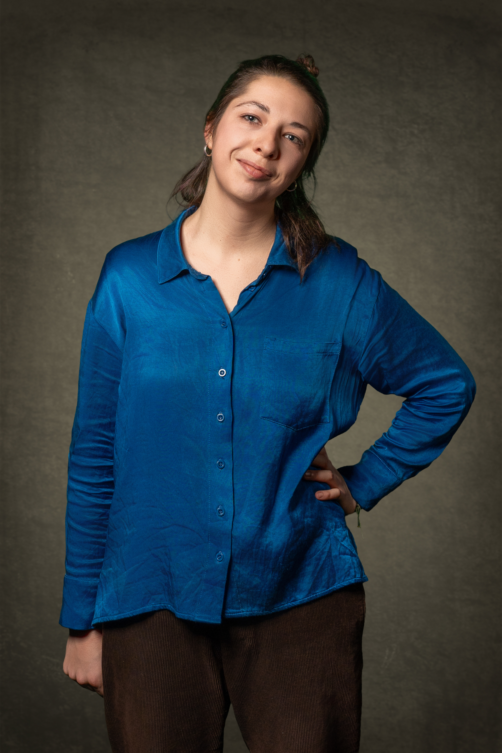 Person steht mit einer Hand in der Hüfte und trägt eine blaue Bluse und braune Hose