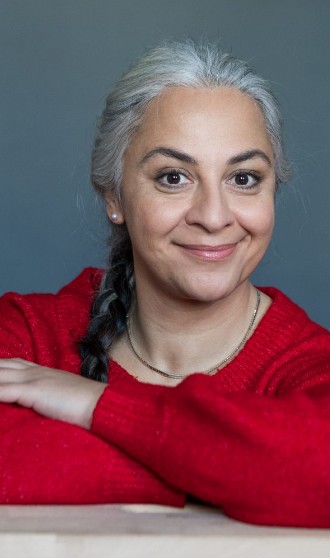 Porträt einer lächelnden Frau mit grauen Haaren in rotem Pullover vor grauem Hintergrund.