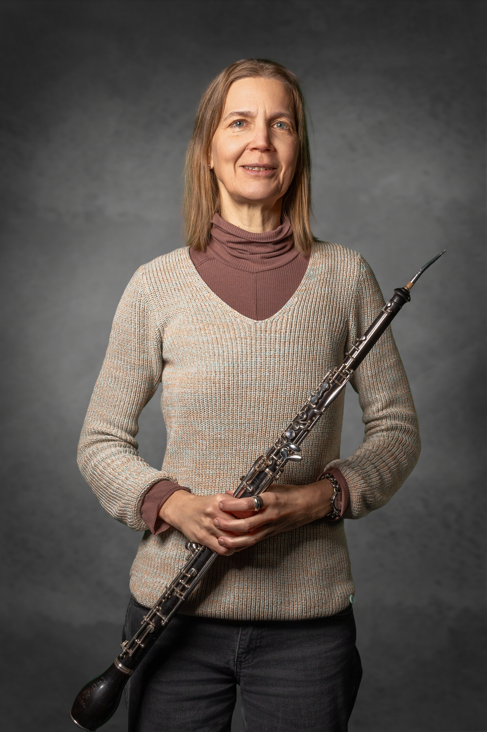 Sängerin mit Oboe in der Hand, lächelnd, in beigem Pullover, vor grauem Hintergrund.