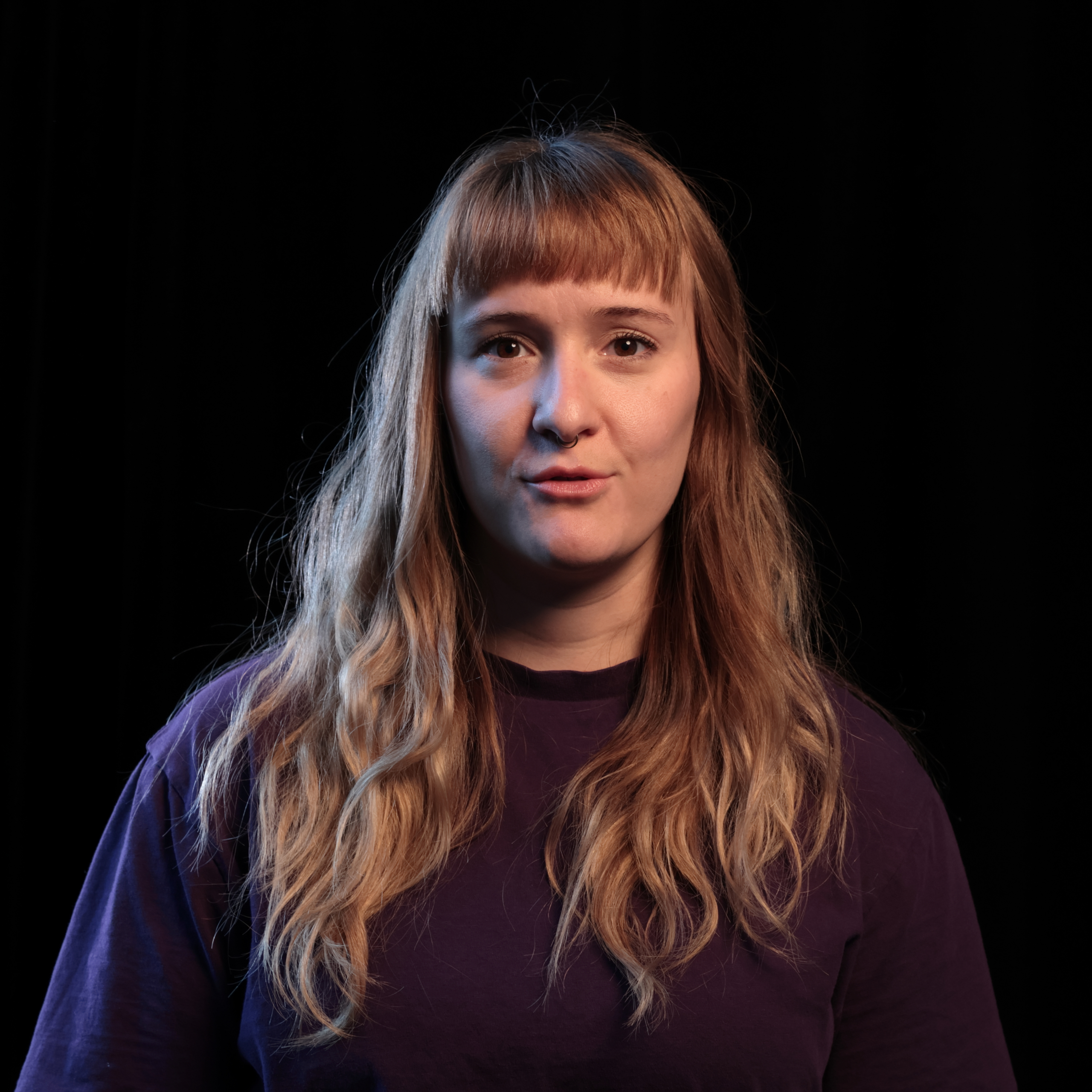 Person mit langen, welligen, blonden Haaren trägt ein dunkles T-Shirt vor schwarzem Hintergrund.