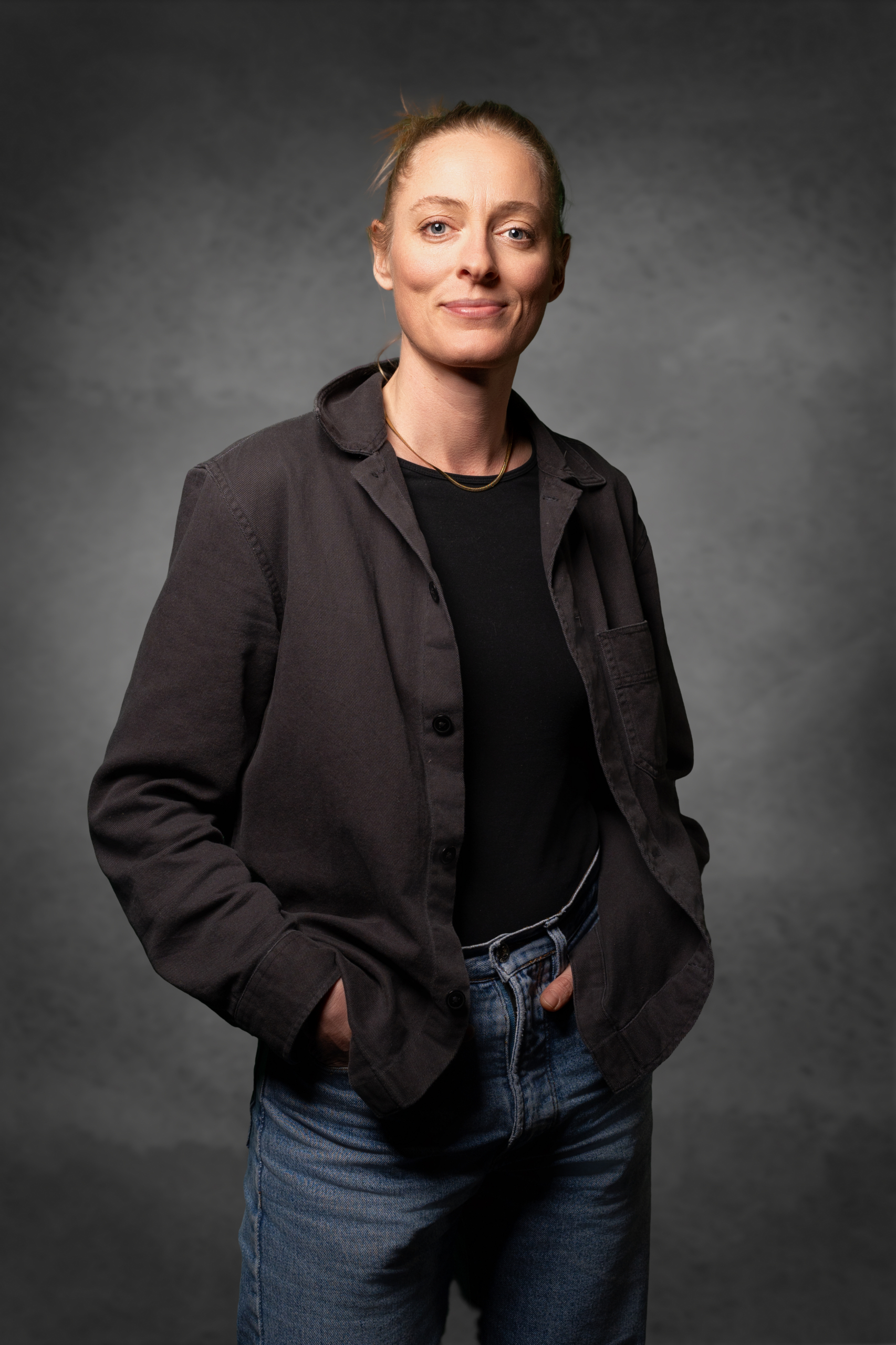 Porträt einer Frau in schwarzem Oberteil und Jacke mit Jeans, lächelnd vor grauem Hintergrund.