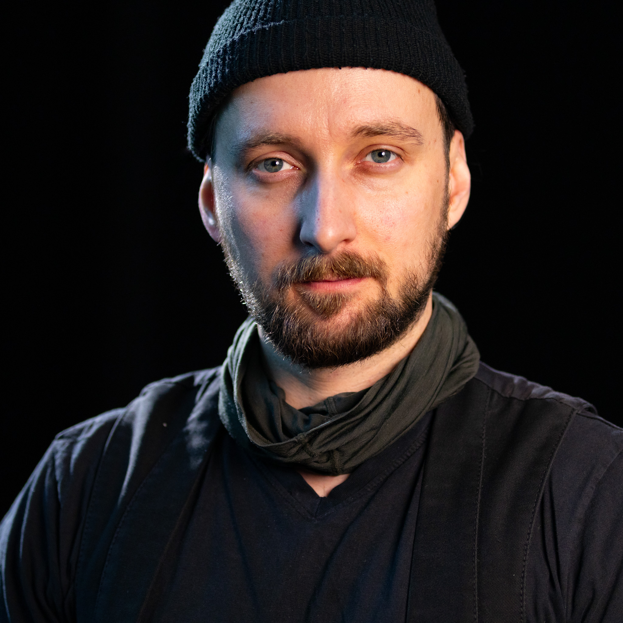 Portrait eines Mannes mit Bart und Mütze, schwarze Kleidung und Schal, stehend vor dunklem Hintergrund.