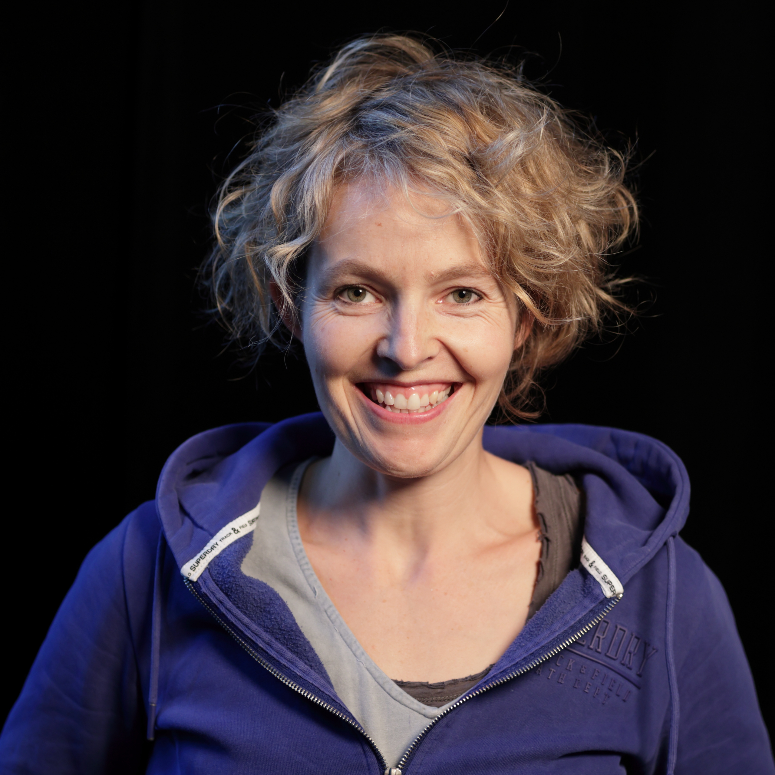 Frau mit blonden lockigen Haaren und blauem Kapuzenpullover vor schwarzem Hintergrund.