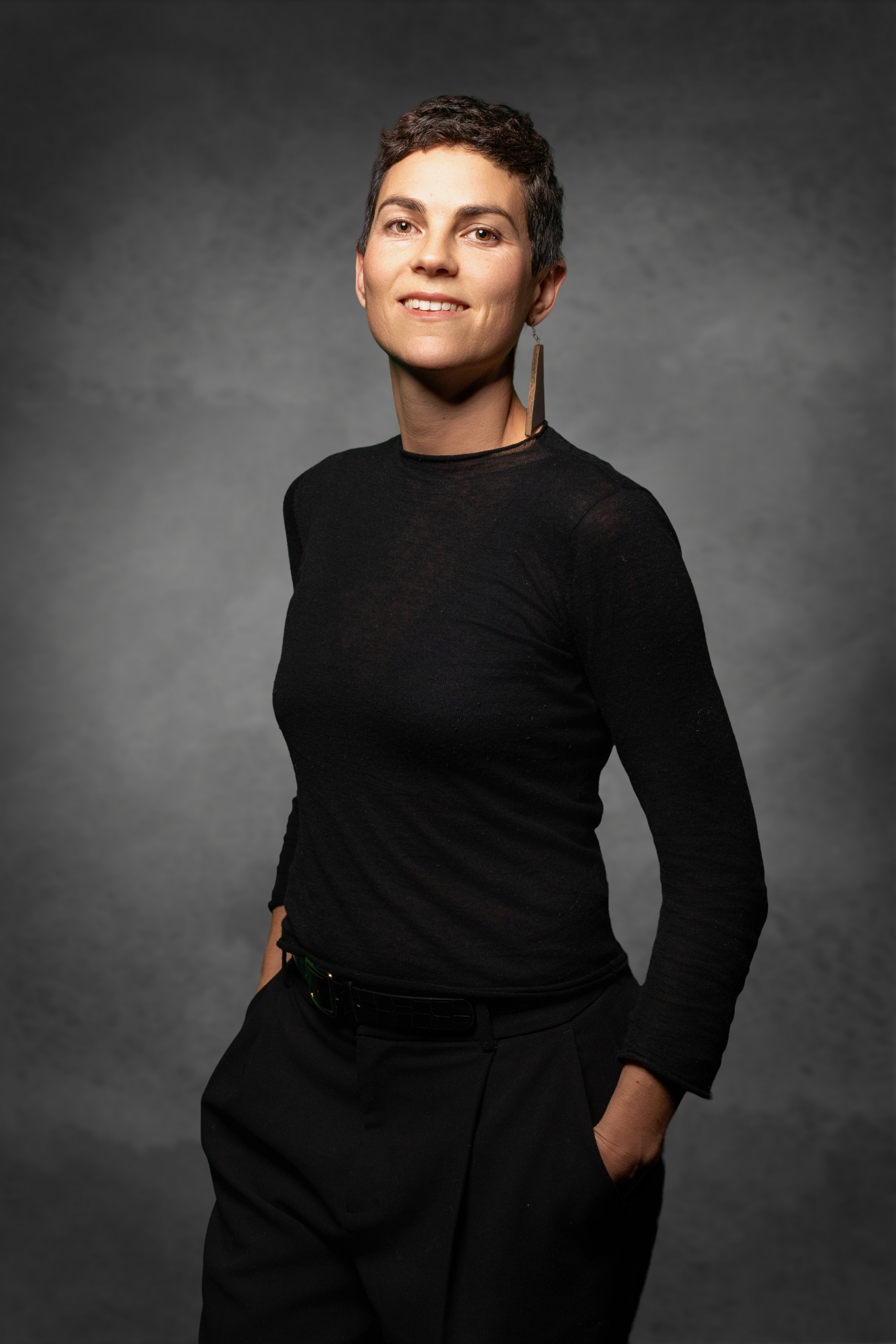 Porträt einer Frau mit kurzen Haaren, schwarzem Shirt und eleganter Pose, in einem grauen Hintergrund.