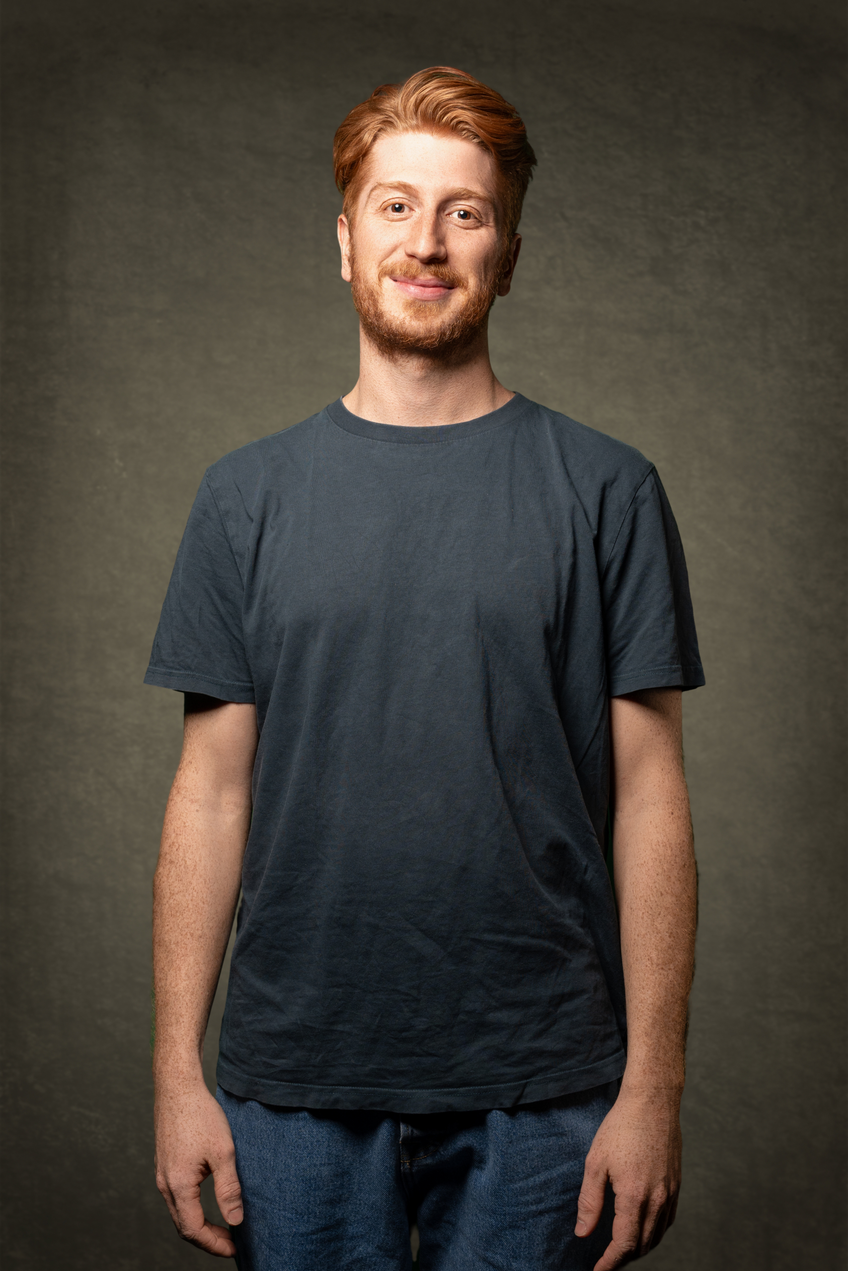 Mann mit rotem Haar in schlichtem dunkelblauen T-Shirt und Jeans vor neutralem Hintergrund.