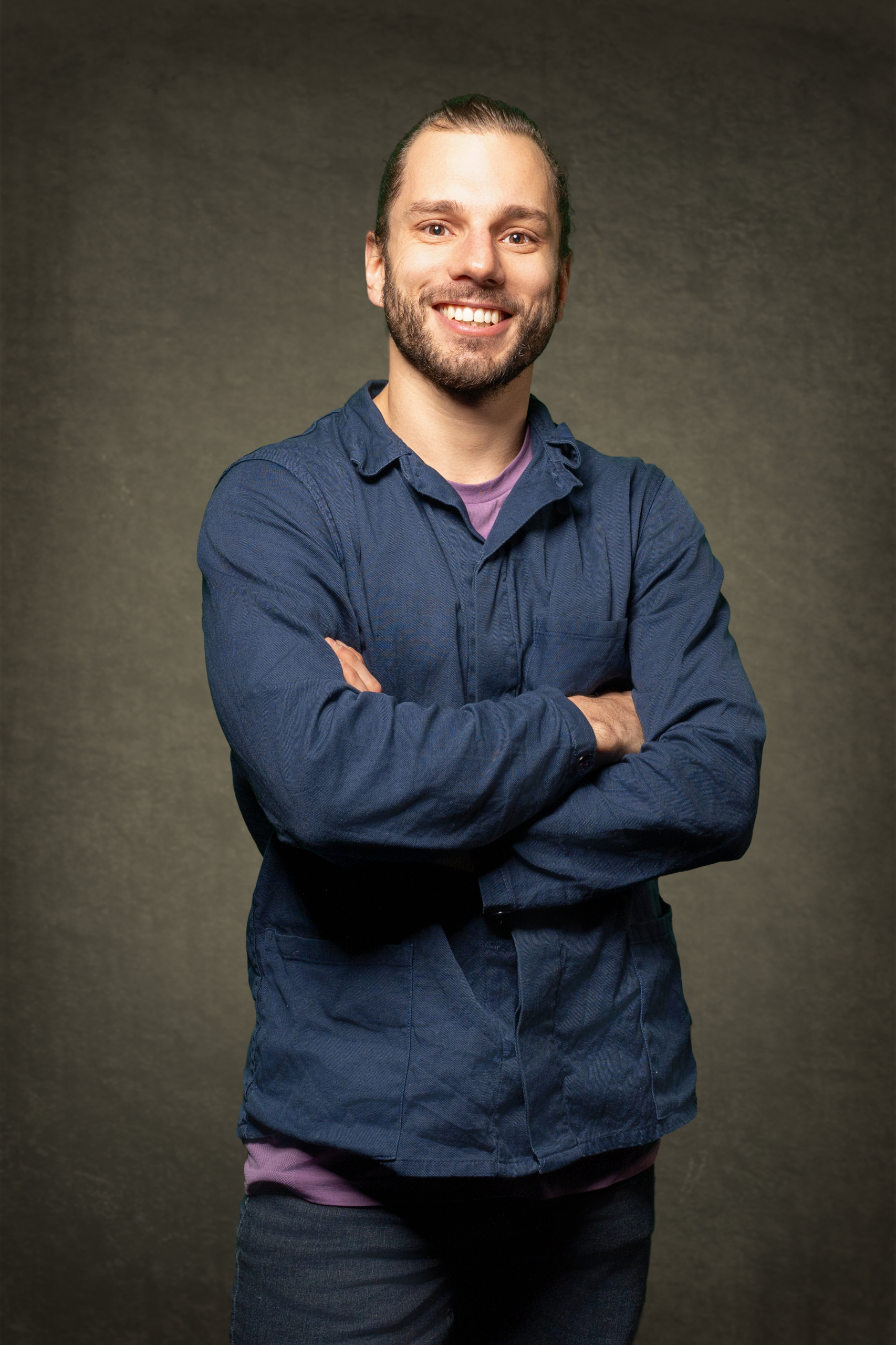 Lächelnder Mann mit kurzen Haaren, trägt eine blaue Jacke, steht mit verschränkten Armen vor einem neutralen Hintergrund.