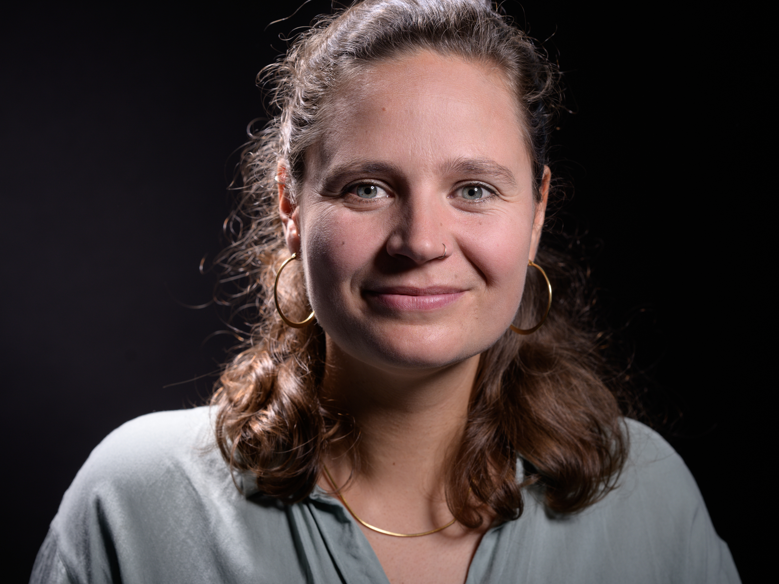 Porträt einer lächelnden Frau mit lockigem Haar, goldenen Ohrringen und einem grauen Oberteil.