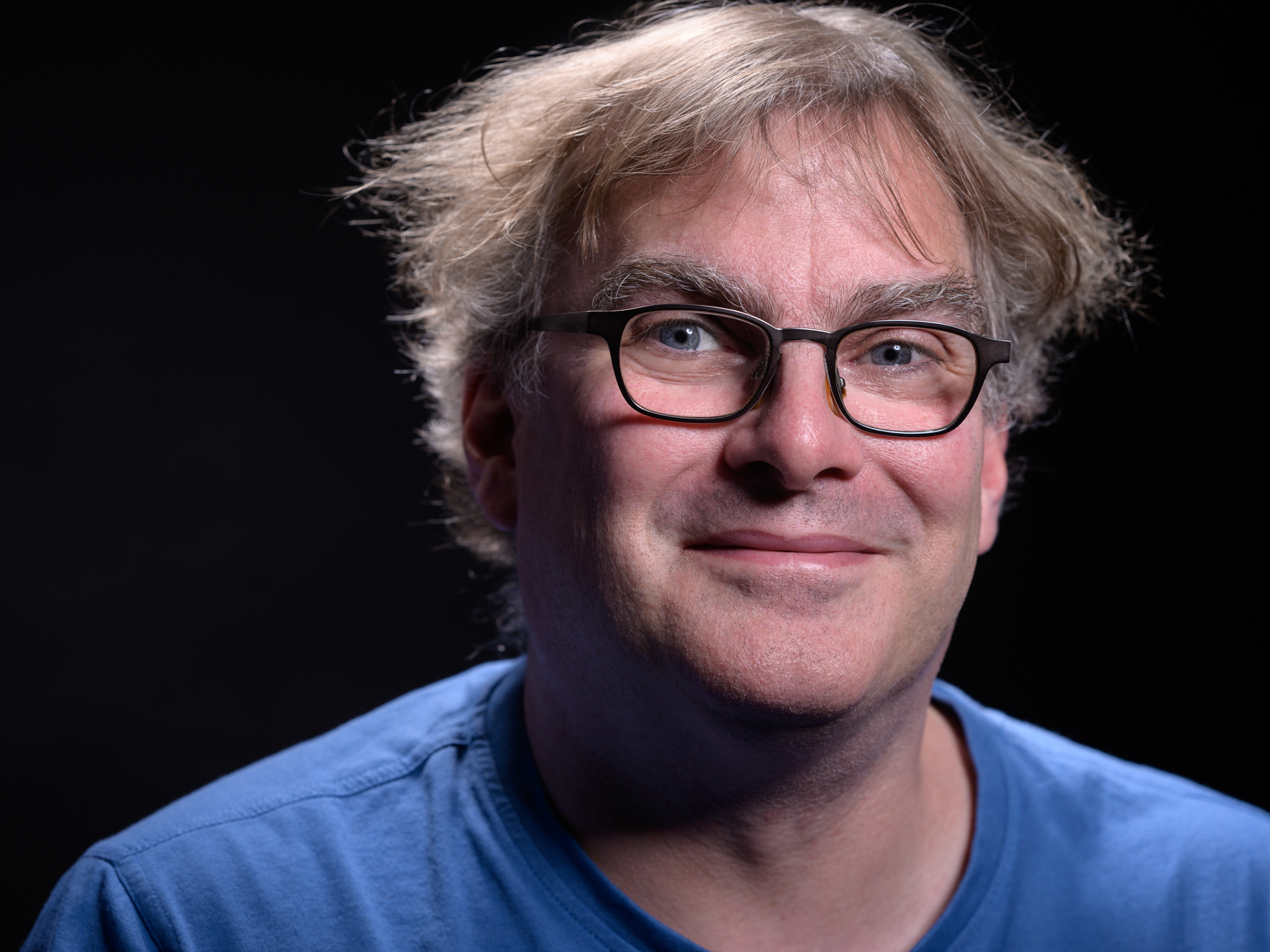 Oberkörper eines Mannes mit blonden, leicht zerzausten Haaren, der ein blaues T-Shirt trägt, vor dunklem Hintergrund.