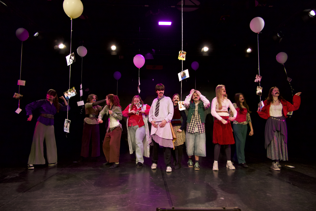 Eine Gruppe von Jugendlichen steht auf einer Bühne und schaut in unterschiedliche Richtungen. An der Decke hängen Luftballons mit Fotos an Schnüren.