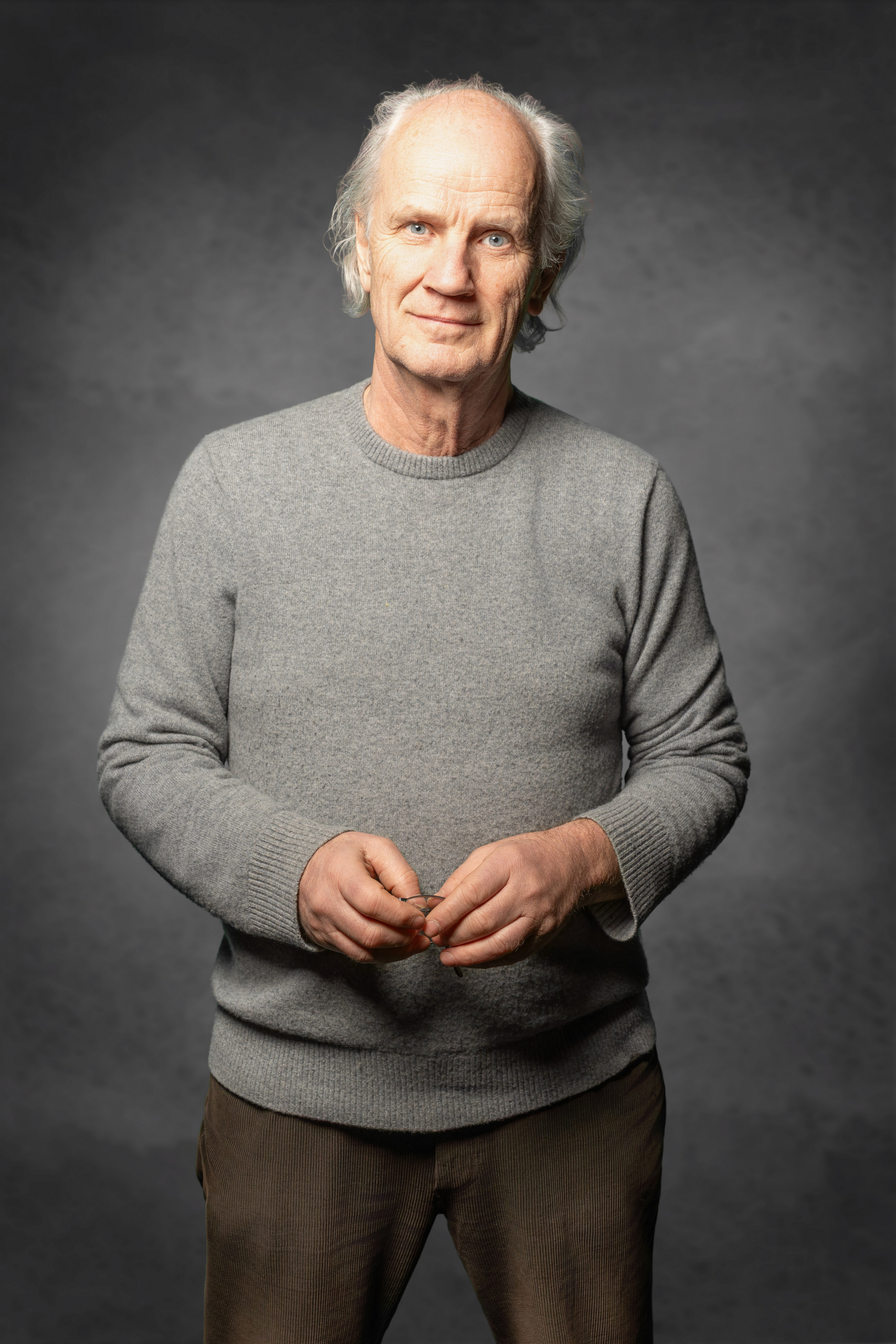 Älterer Mann mit grauem Pullover und brauner Hose steht vor dunklem Hintergrund und hält ein kleines Objekt in den Händen