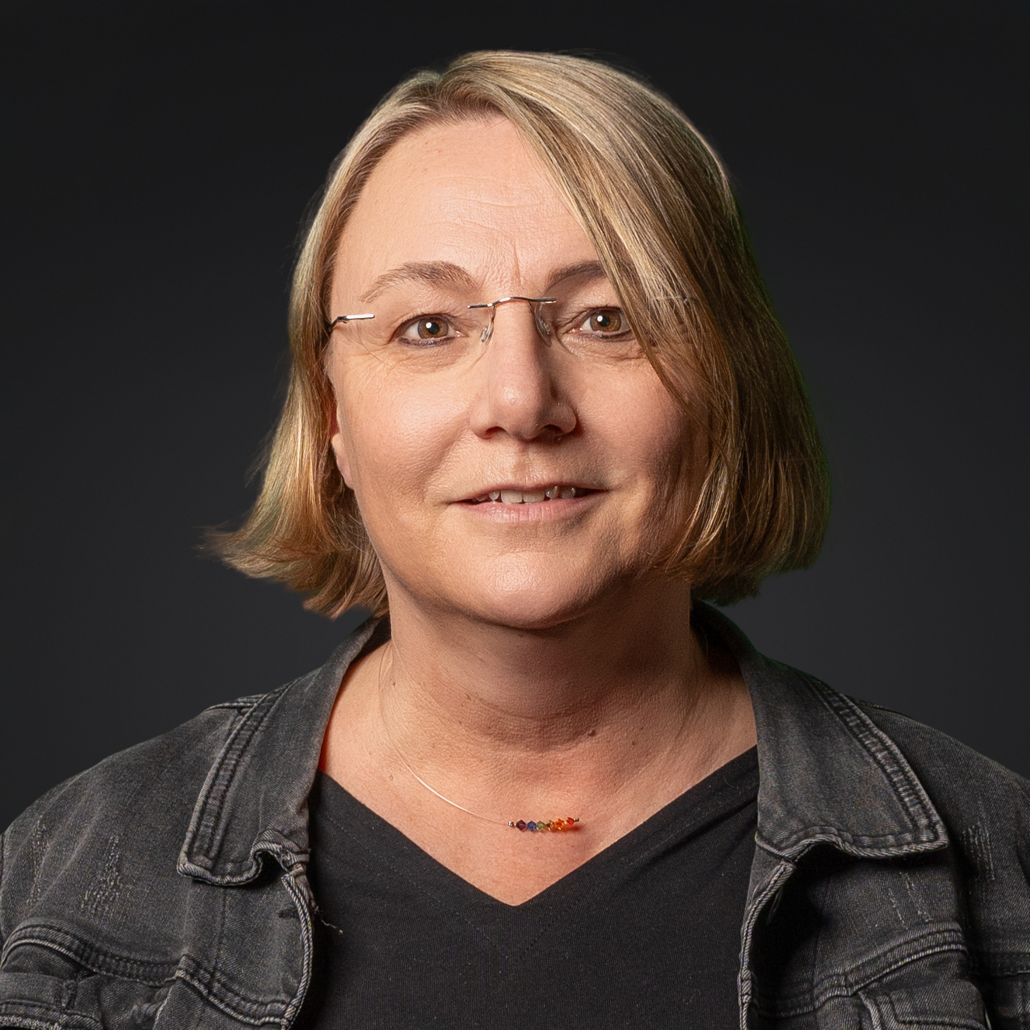 Oberkörper einer Person mit schulterlangen blonden Haaren, schwarzem T-Shirt, Jeansjacke und einer Kette mit bunten Perlen.