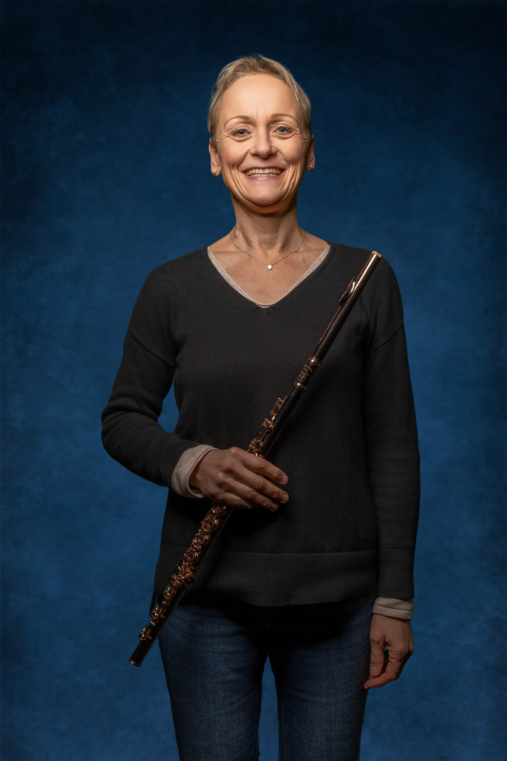 Portrait einer lächelnden Frau mit Flöte vor blauem Hintergrund, in lässigem Outfit.