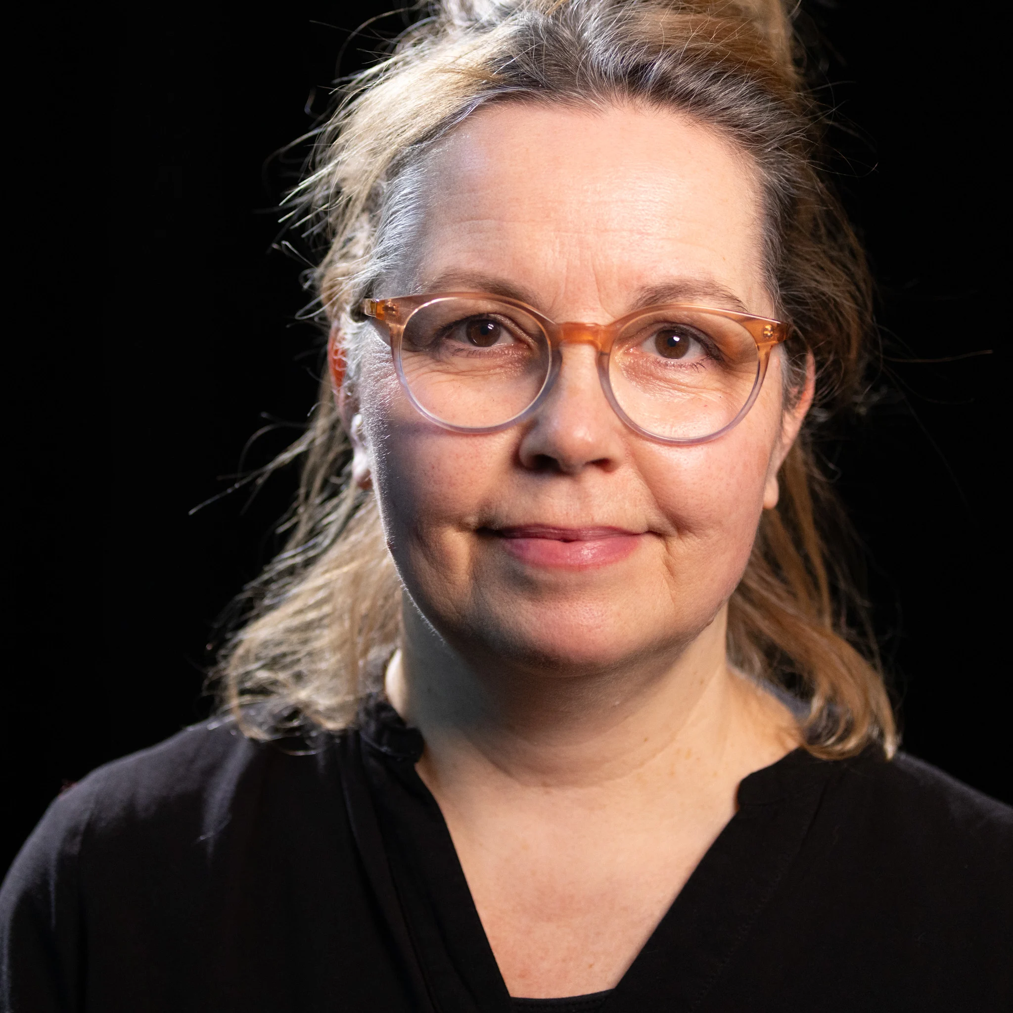 Person mit schulterlangem, hellbraunem Haar, das locker zurückgebunden ist, trägt ein schwarzes Oberteil mit V-Ausschnitt vor schwarzem Hintergrund.