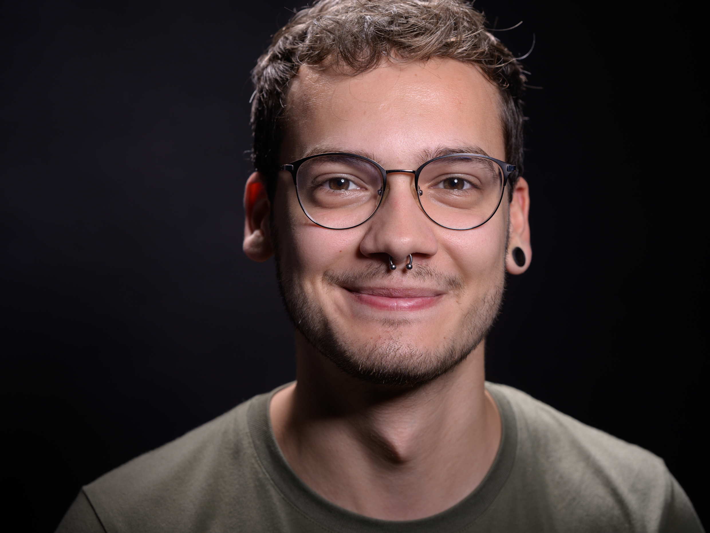 Oberkörper eines Mannes mit kurzem, gewelltem Haar, olivgrünem T-Shirt vor schwarzem Hintergrund.