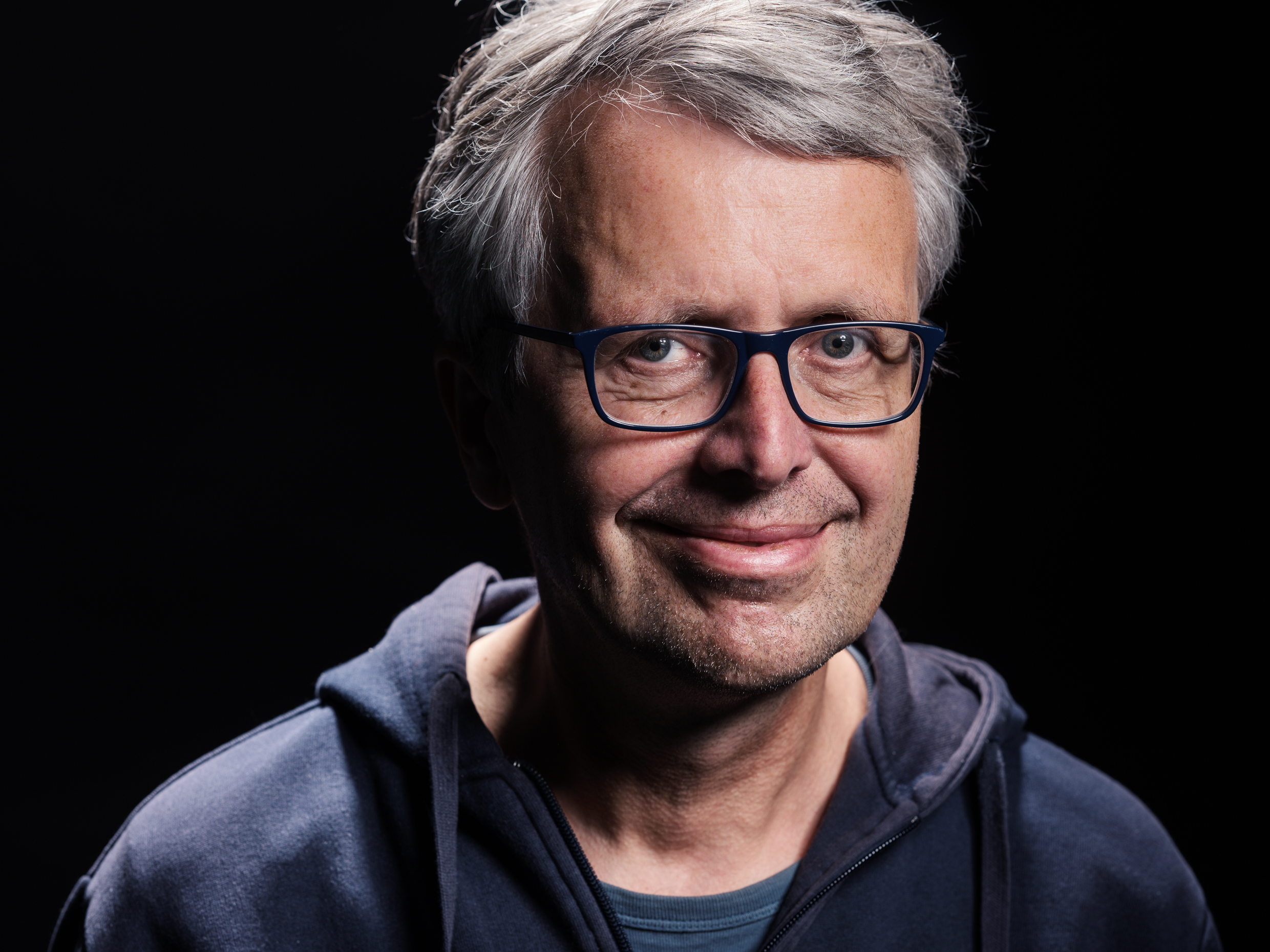 Mann mit grauen Haaren trägt dunkelblaue Kapuzenjacke und dunkles T-Shirt vor schwarzem Hintergrund.