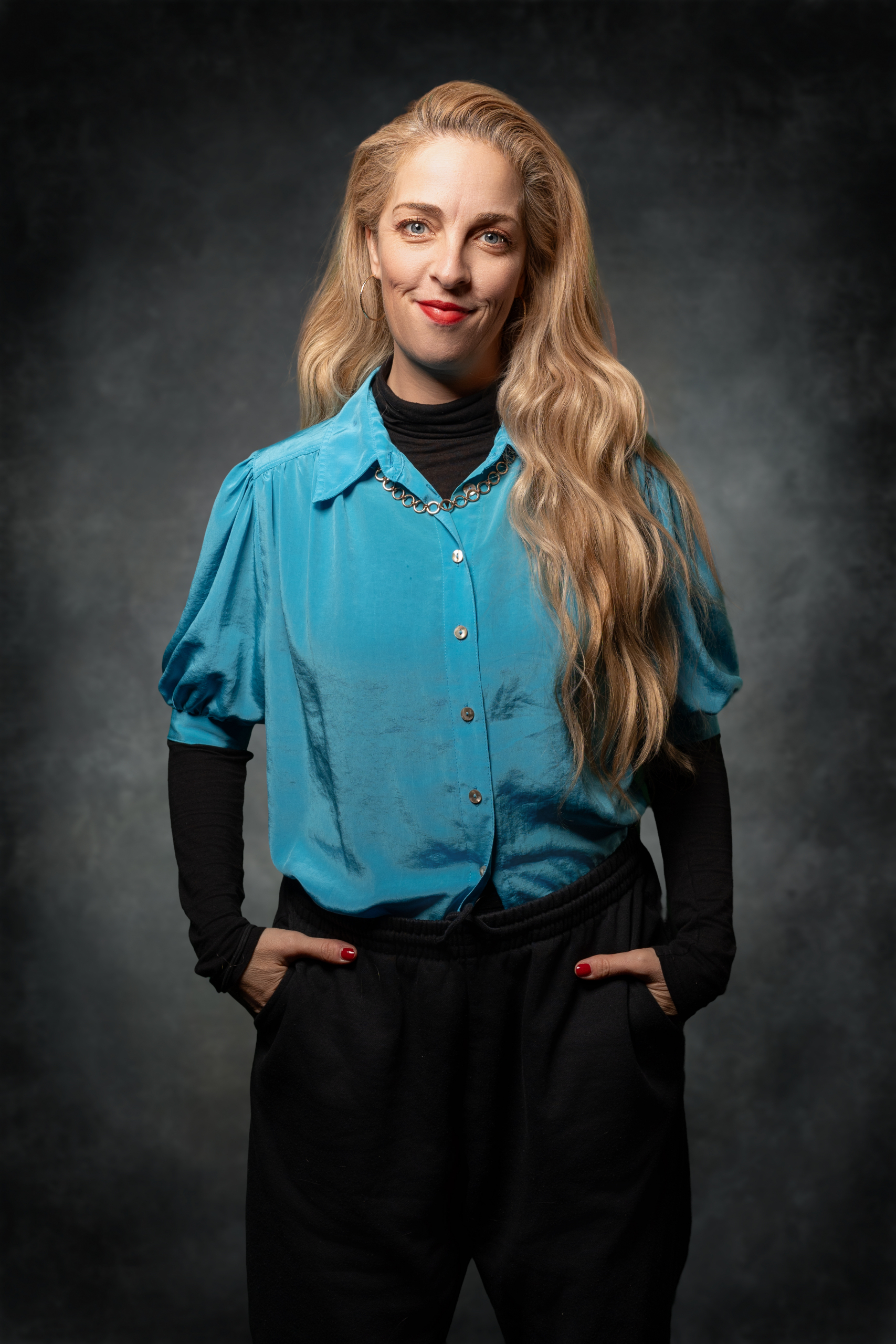 Frau mit langen, blonden Haaren in einer blauen Bluse und schwarzen Hosen, posiert vor dunklem Hintergrund.