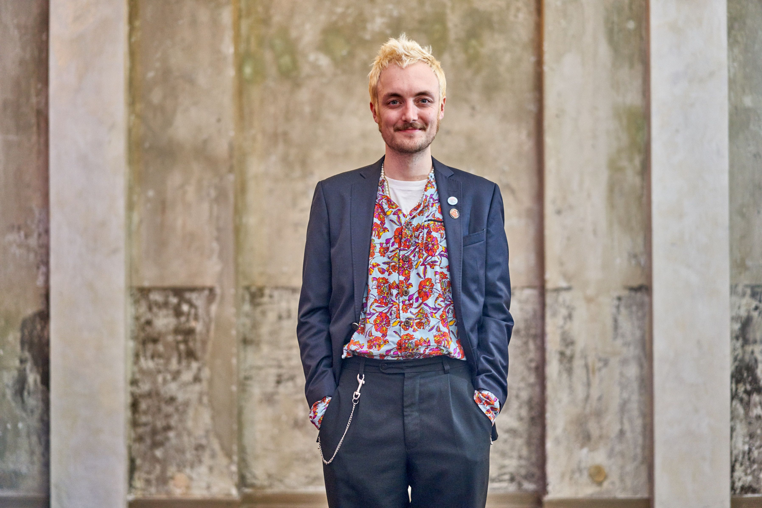 Person mit kurzem blonden Haar trägt ein gemustertes Hemd unter einem dunklen Sakko und eine Kette an der Hose vor einer Wand mit vertikalen Strukturen.