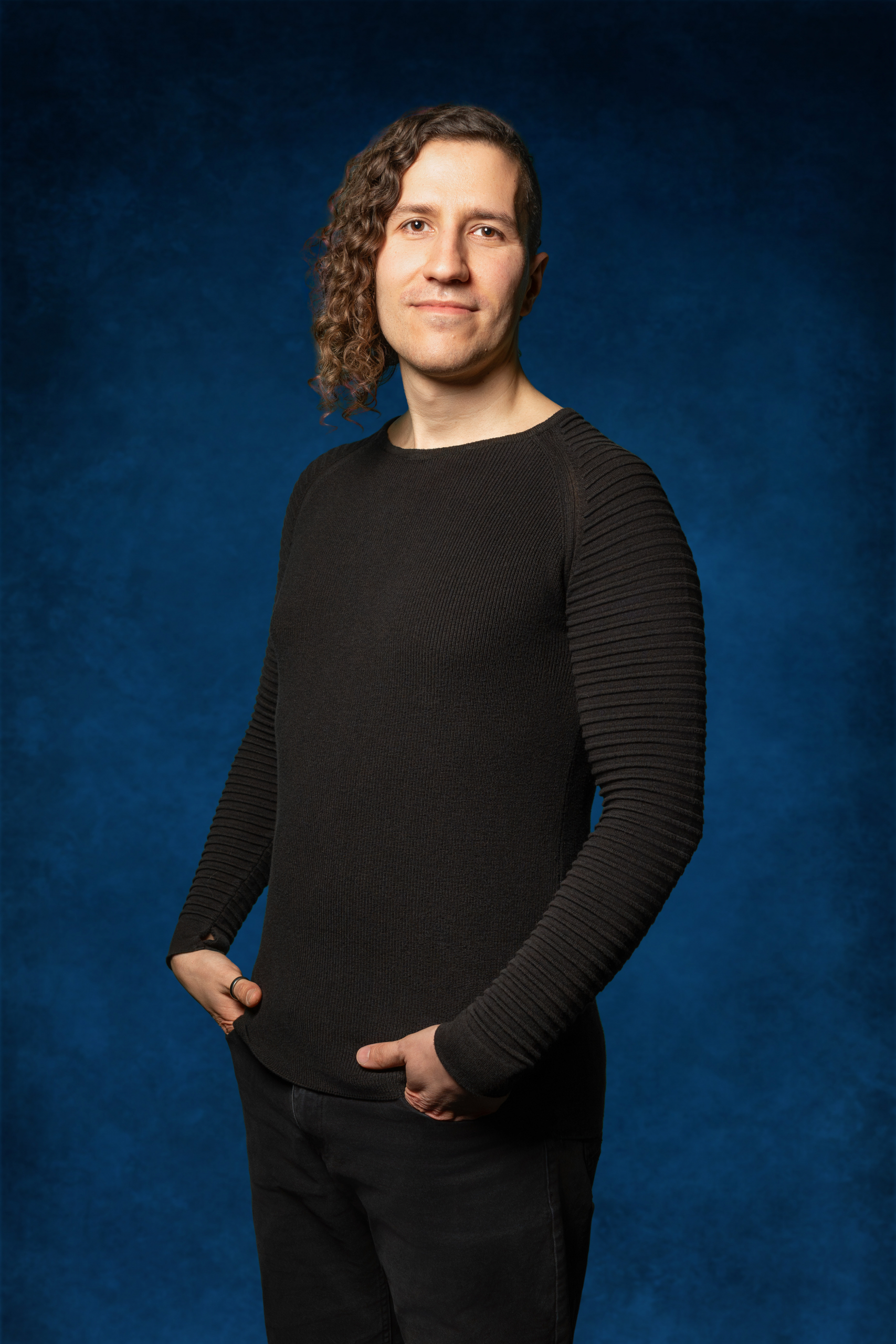 Person mit lockigem Haar, die schwarze, gerippte Langarmjacke und schwarze Hose trägt und die Hände in die Hosentaschen steckt vor blauem Hintergrund