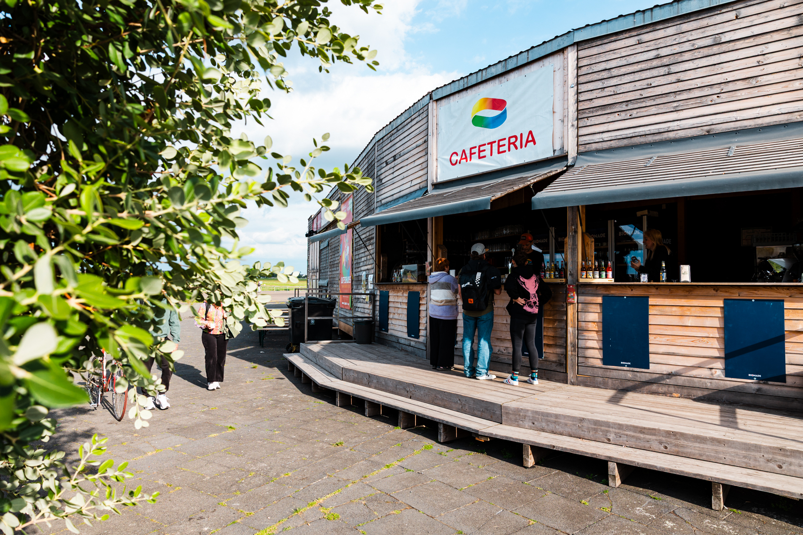 Holzgebäude mit offenem Fenster und Schild 'CAFETERIA', davor stehen mehrere Menschen in einer Reihe.