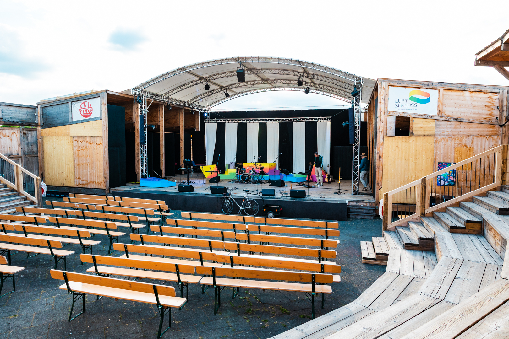 Freiluftbühne mit überdachtem Bühnenbereich, davor mehrere Reihen hölzerner Sitzbänke und rechts hölzerne Treppenstufen