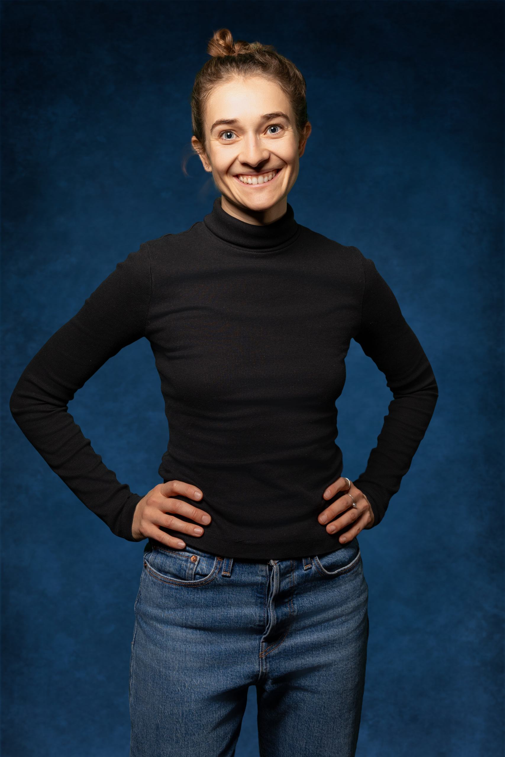 Lächelnde Frau in schwarzem Rollkragenpullover und Jeans präsentiert sich vor blauem Hintergrund.