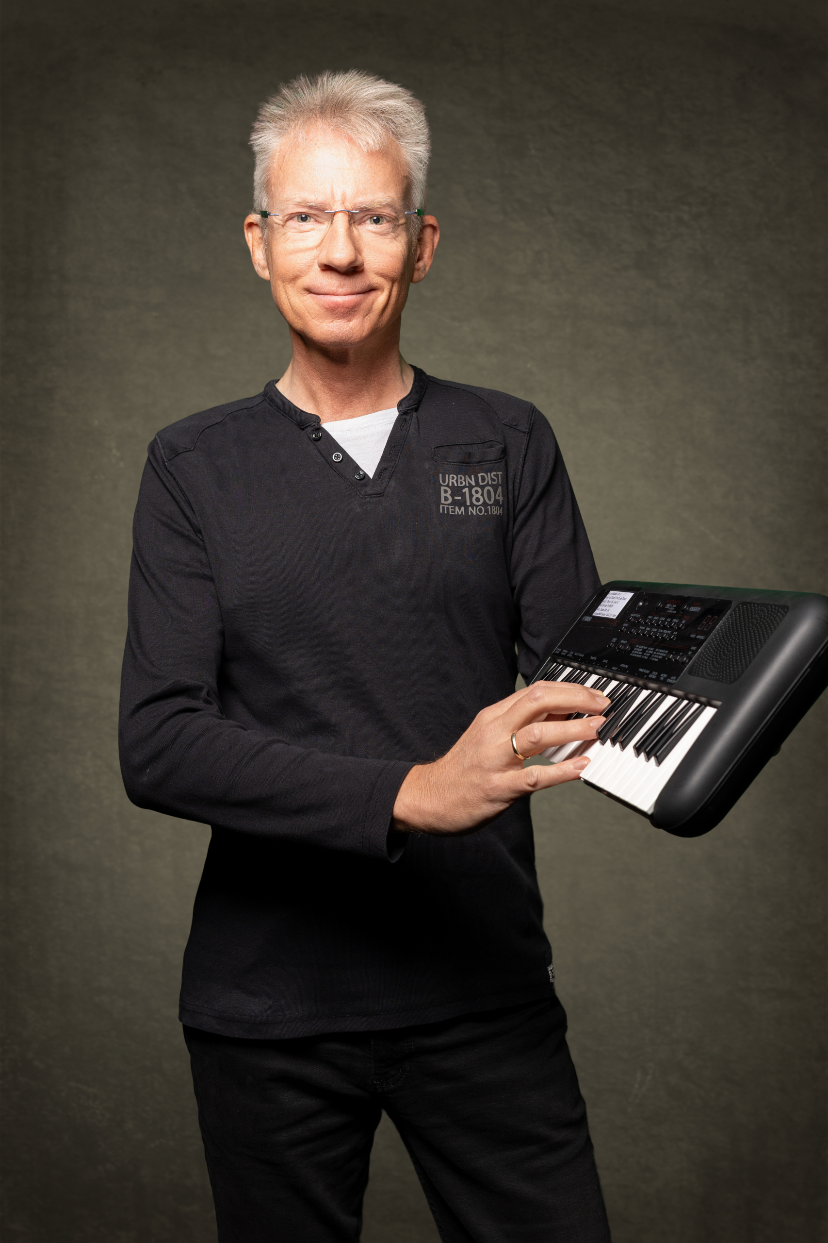 Mann mit Brille hält ein Keyboard, lächelt in einem grauen Raum. Keyboard-Modell nicht angegeben.