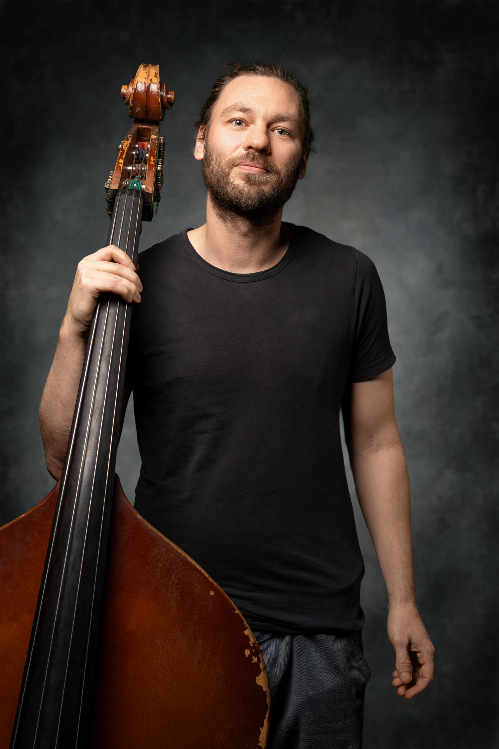 Mann mit einem Kontrabass in der Hand, lächelnd, trägt ein schwarzes T-Shirt und hat einen lässigen Look.