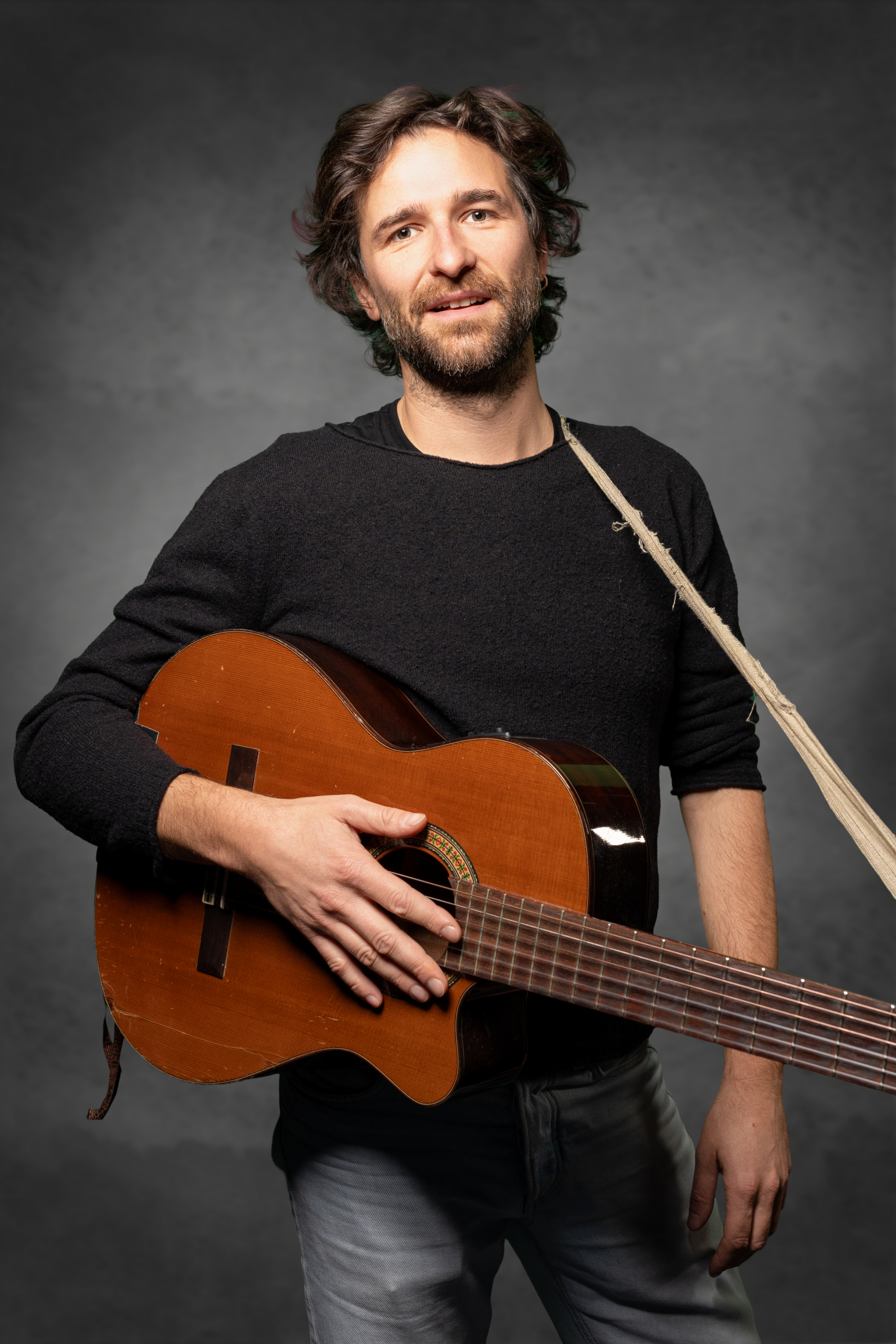 Mann mit Gitarre in der Hand, trägt einen schwarzen Pullover und hat eine entspannte Ausstrahlung.