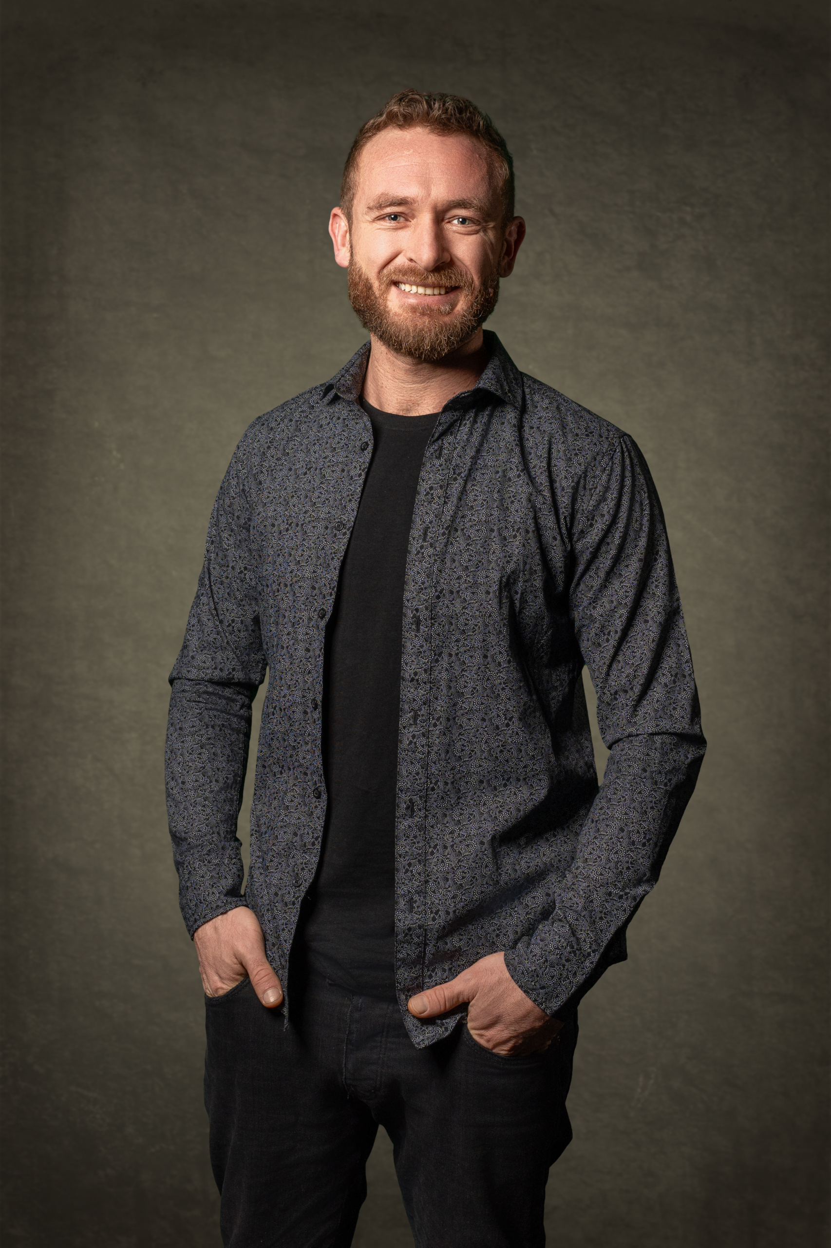 Mann in dunkler, gepunkteter Hemdjacke mit schwarzem T-Shirt, lächelt freundlich vor grauem Hintergrund.