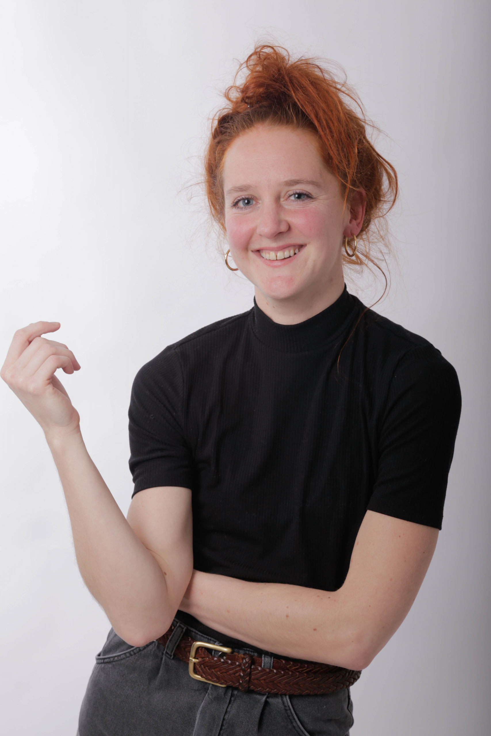 Junge Frau mit rotem Haar und schwarzem Shirt, lächelt und zeigt eine Geste vor einem neutralen Hintergrund.