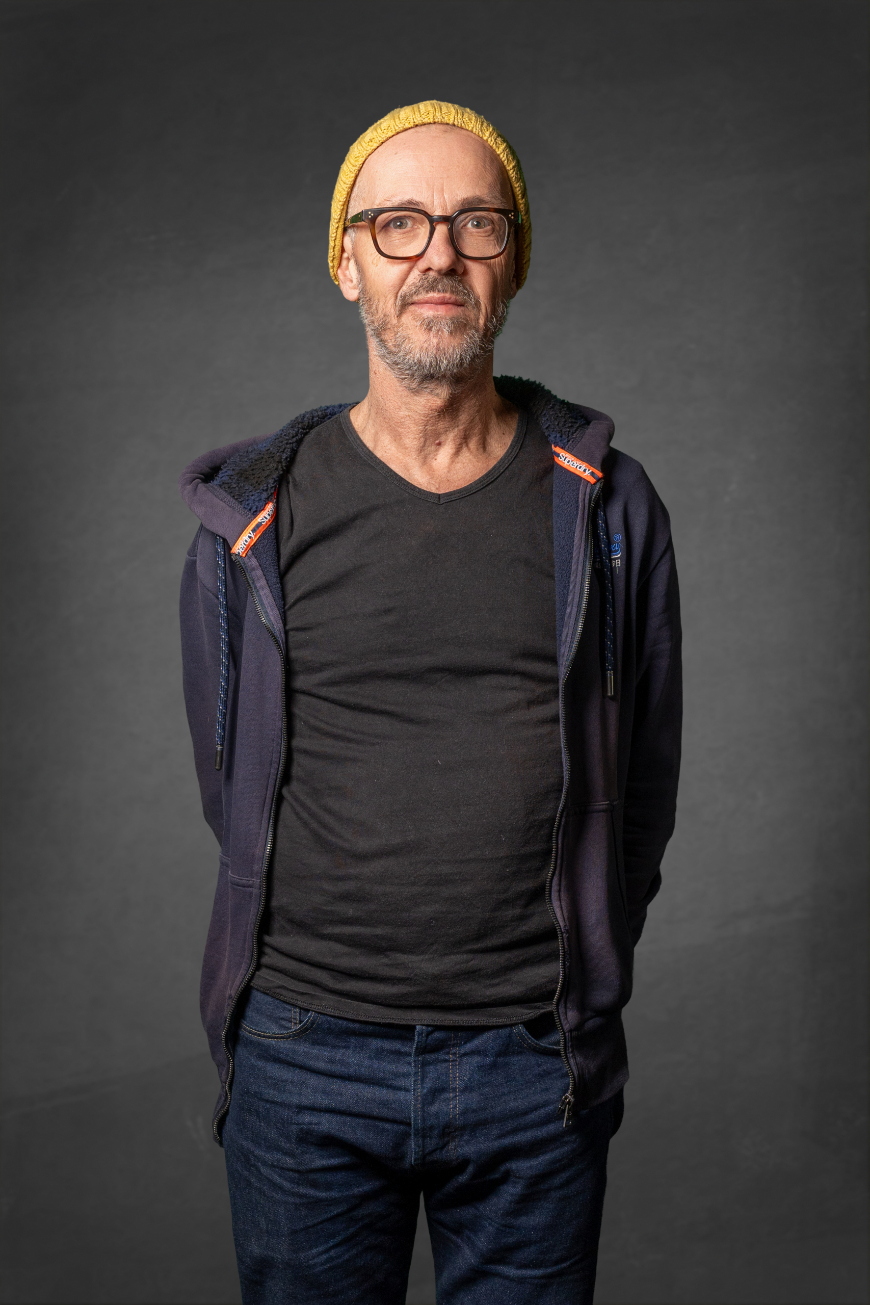 Porträt eines Mannes mit Brille, gelbem Hut und schwarzem T-Shirt, stehend in neutraler Pose.