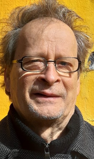 Porträt eines älteren Mannes mit Brille vor einer gelben Wand.