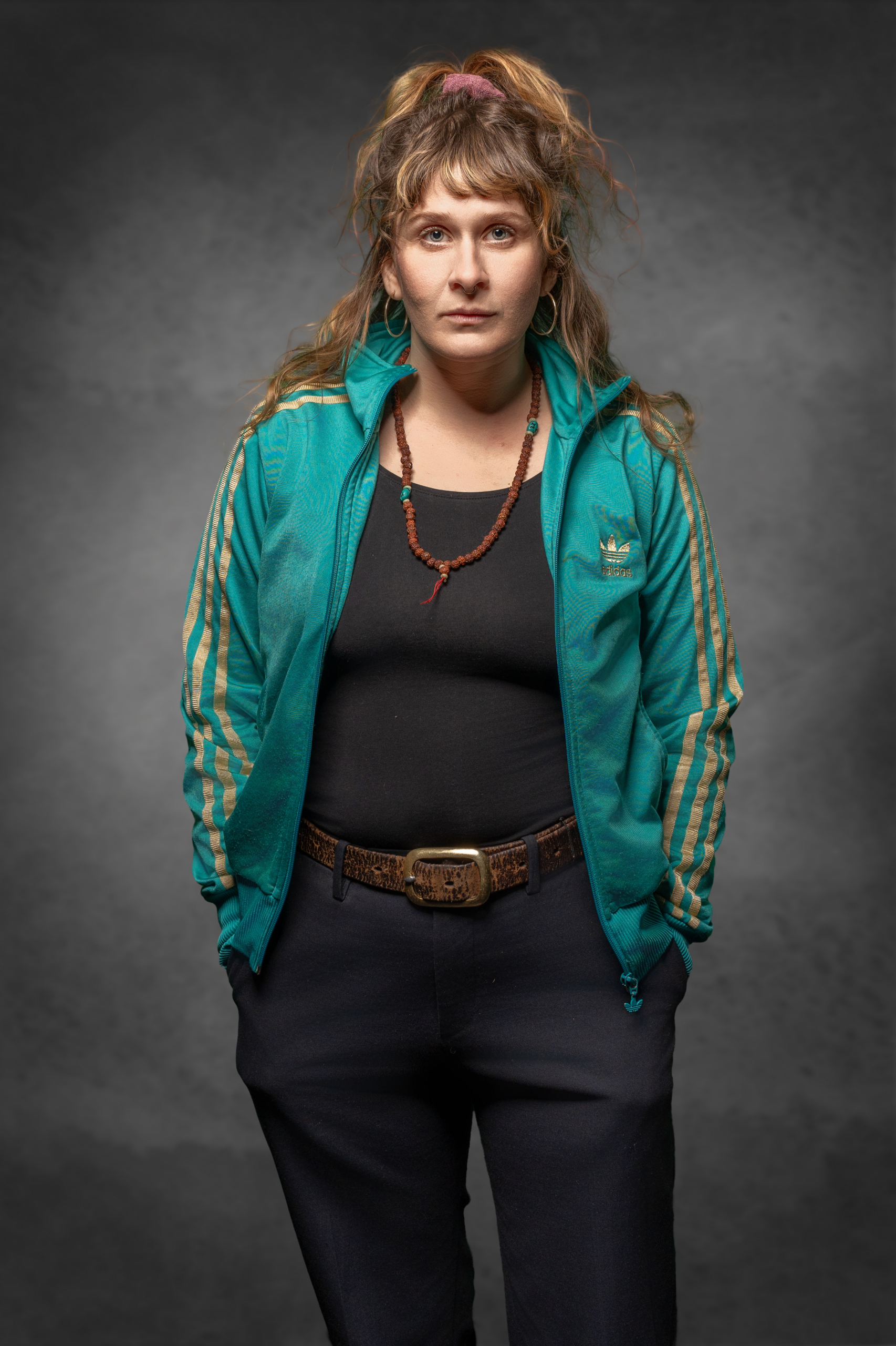 Frau in teal Adidas-Jacke mit goldenen Streifen, schwarzem Oberteil und weiten Hosen, in neutralem Hintergrund.