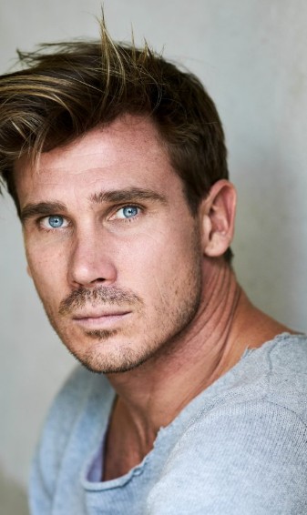 Portrait eines Mannes mit blauen Augen und unordentlichem Haar, trägt ein graues, lässiges Sweatshirt.