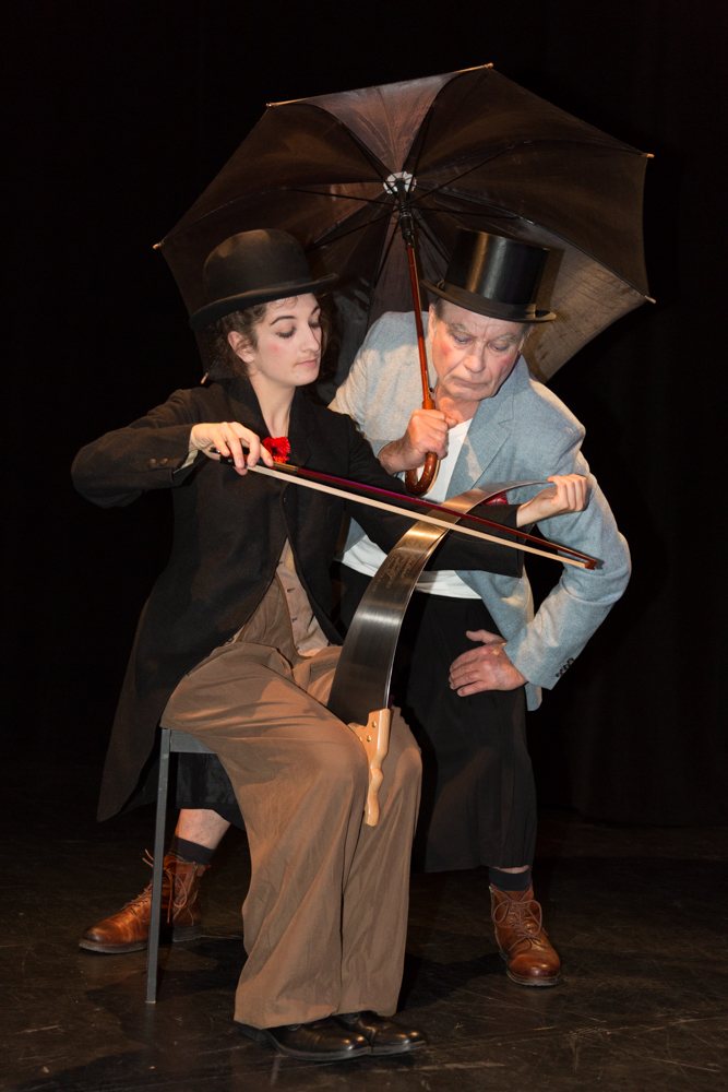 Zwei Personen in schwarzem Zylinder und braunen Schuhen, eine sitzt mit Cello und Bogen, die andere hält einen geöffneten schwarzen Regenschirm über beide.