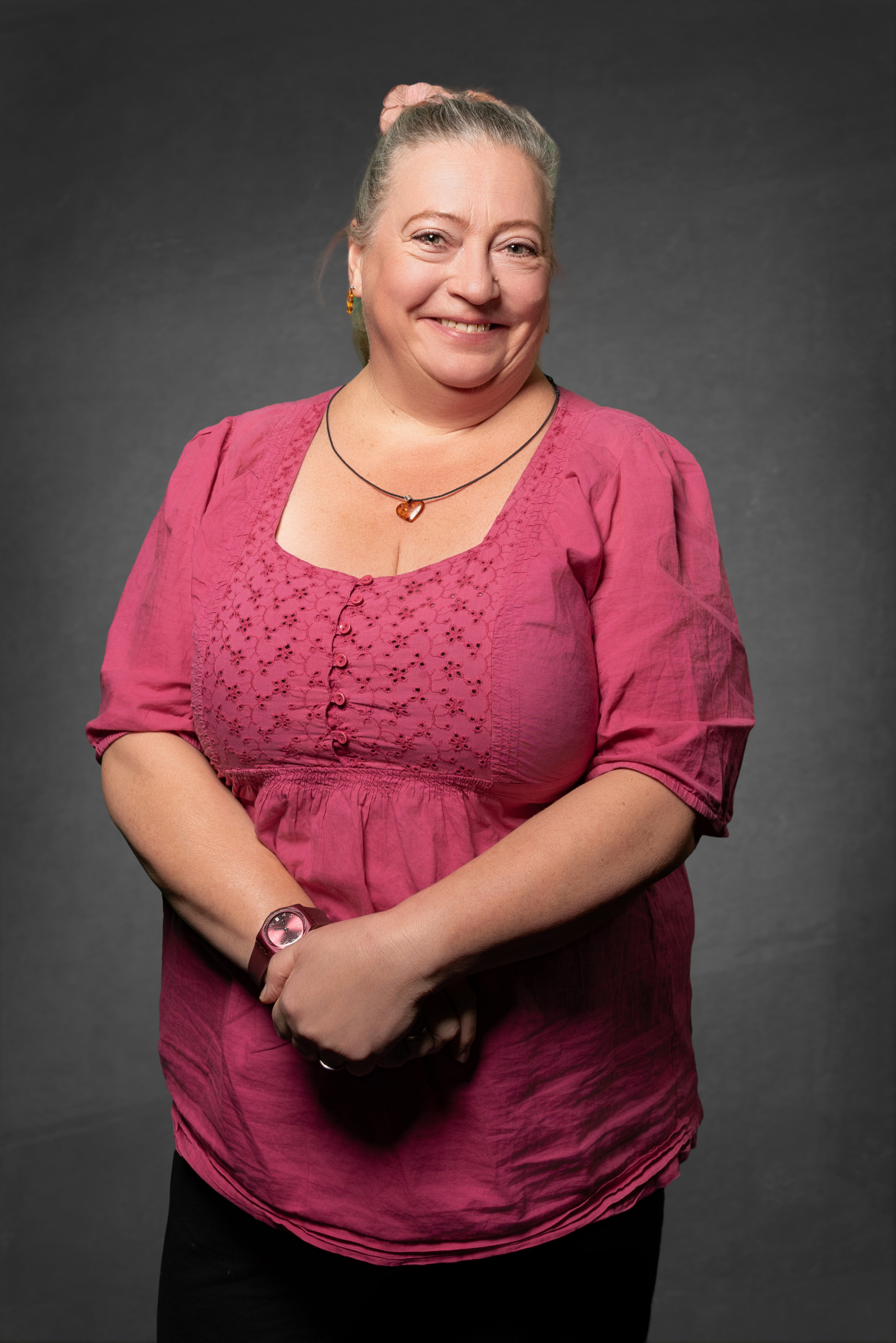 Porträt einer Dame in rosa Bluse mit Knopfdetails, lächelnd vor einem grauen Hinergrund.