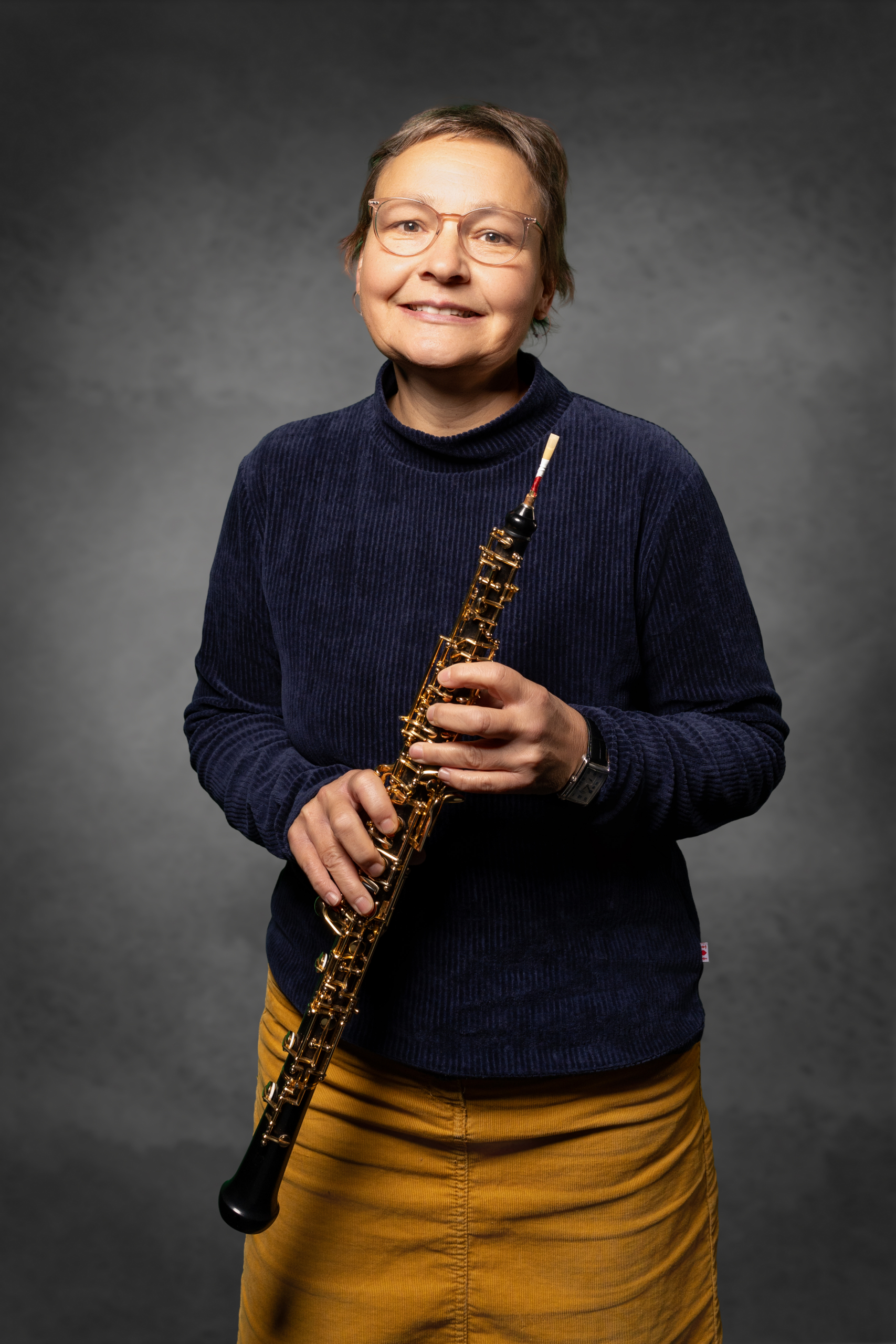 Frau mit Oboe, trägt einen blauen Pullover und einen gelben Rock, lächelt in die Kamera.