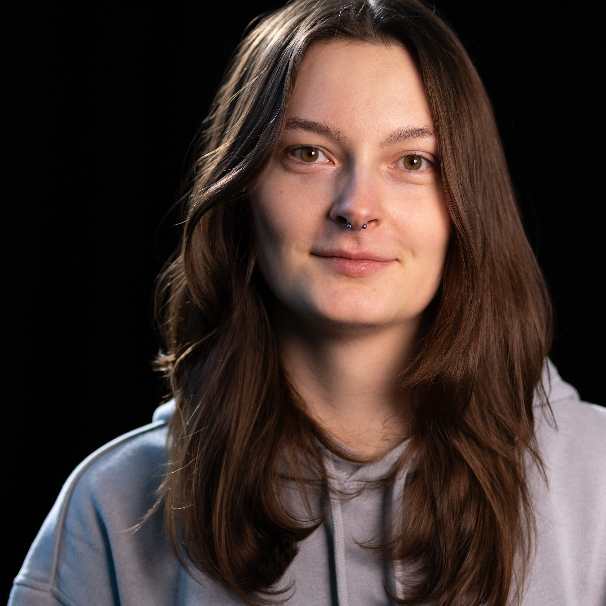 Porträt einer Frau mit langen Haaren und Septum-Piercing in grauem Hoodie vor schwarzem Hintergrund.