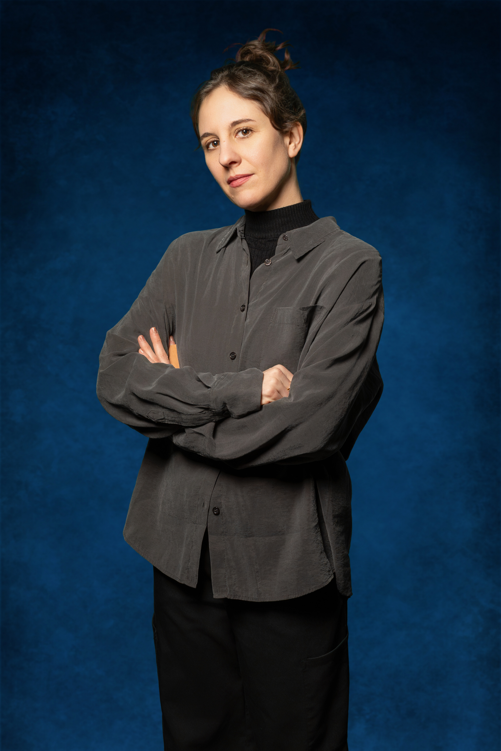 Person mit hochgestecktem Haar trägt ein dunkles Hemd über einem schwarzen Rollkragenpullover und steht mit verschränkten Armen vor blauem Hintergrund