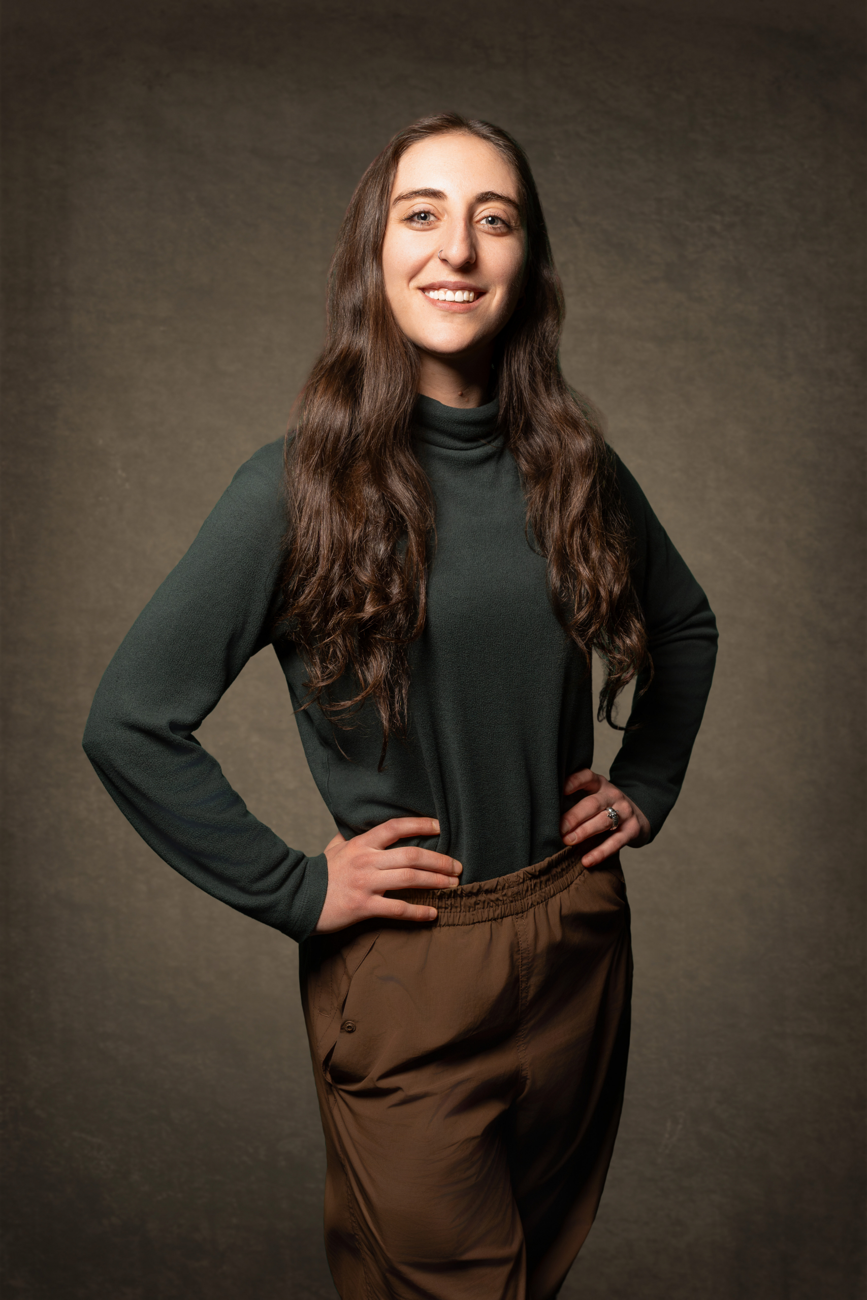 Person mit langen, welligen Haaren trägt einen dunklen Rollkragenpullover und braune Hose und steht mit den Händen in der Hüfte vor einem einfarbigen Hintergrund