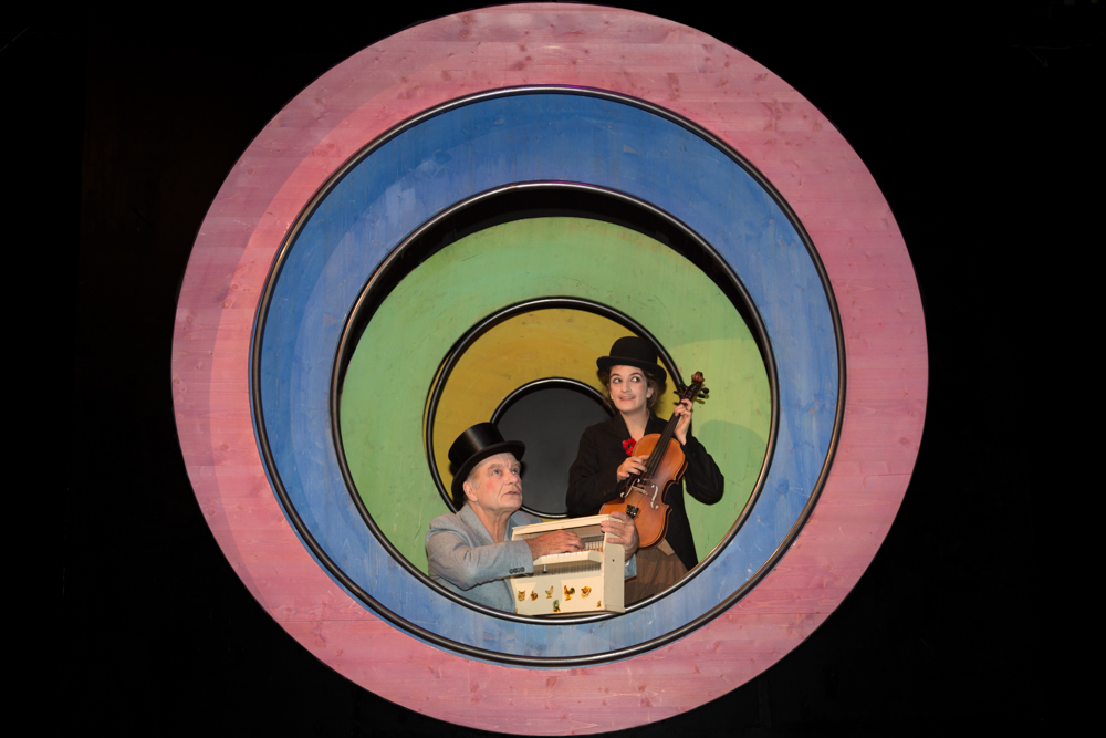 Zwei Personen, eine spielt ein Orgelpositiv, die andere hält eine Geige, umgeben von bunten konzentrischen Kreisen in Rosa, Blau, Grün und Gelb.