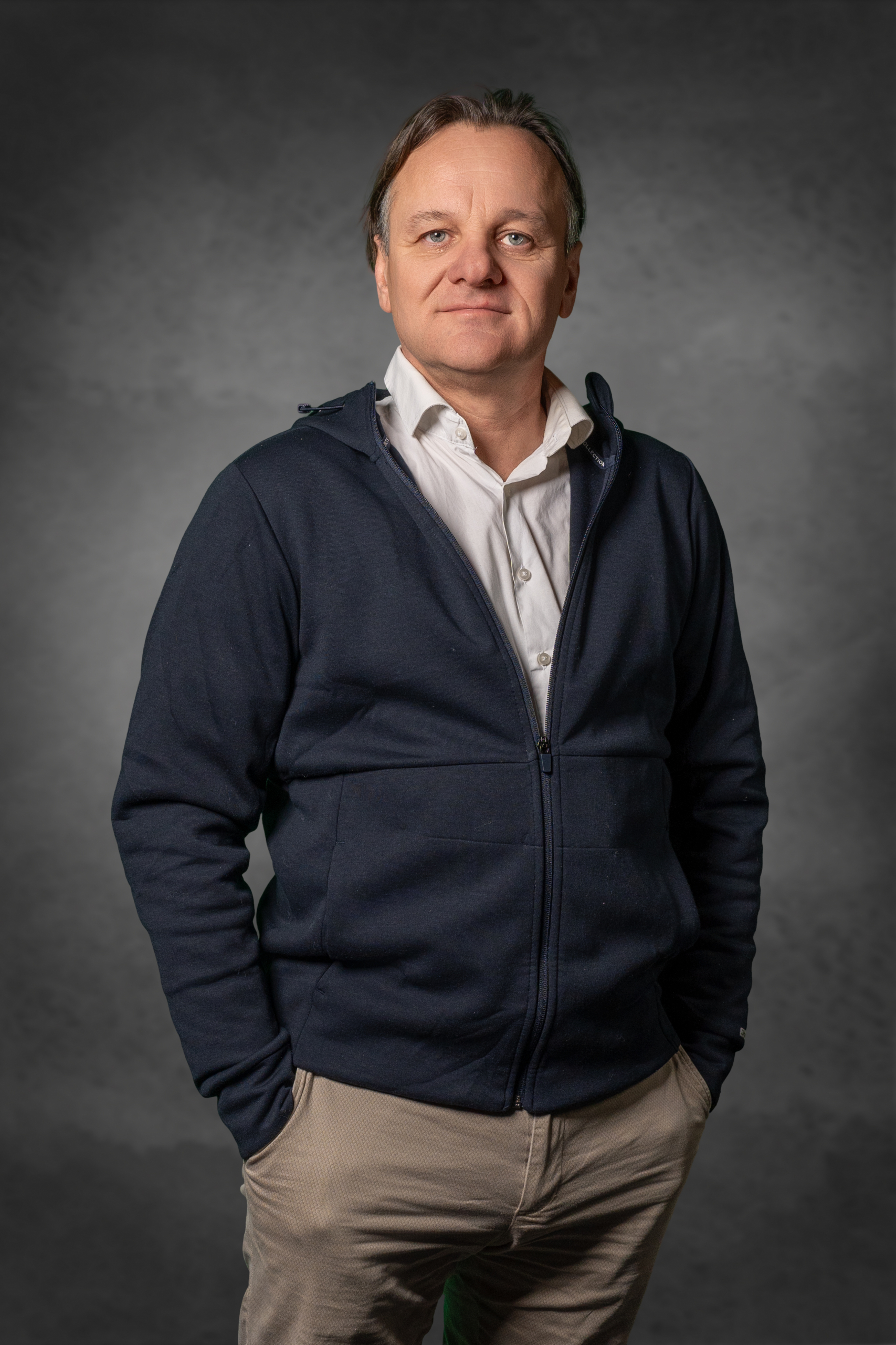 Mann in dunkelblauer Jacke mit Reißverschluss und Hemd vor neutralem Hintergrund.