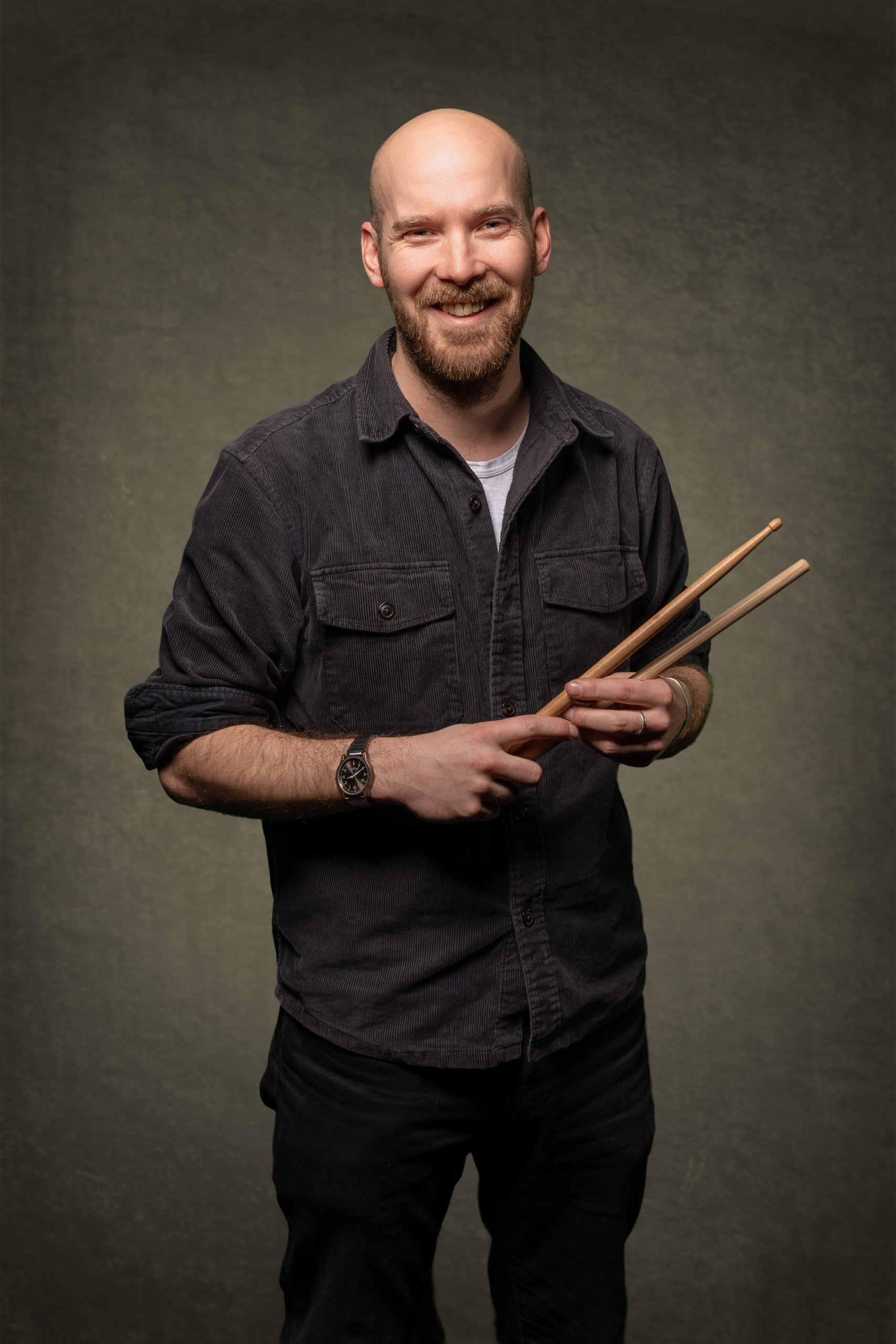 Lächelnder Mann mit Schlagzeugstöcken in der Hand, gekleidet in einer schwarzen Jacke.