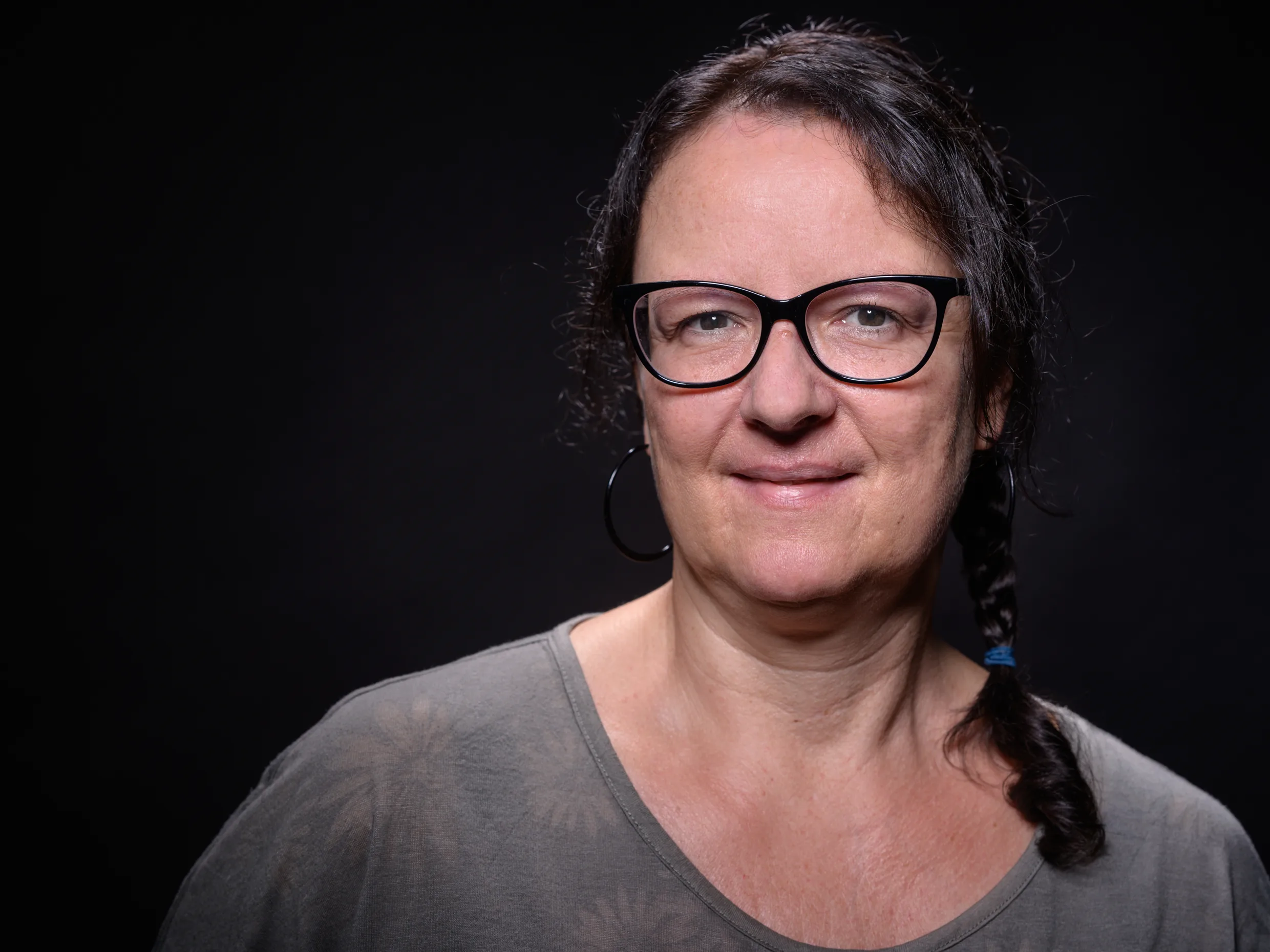 Person mit schulterlangem, hellbraunem Haar, das locker zurückgebunden ist, trägt ein schwarzes Oberteil mit V-Ausschnitt vor schwarzem Hintergrund.