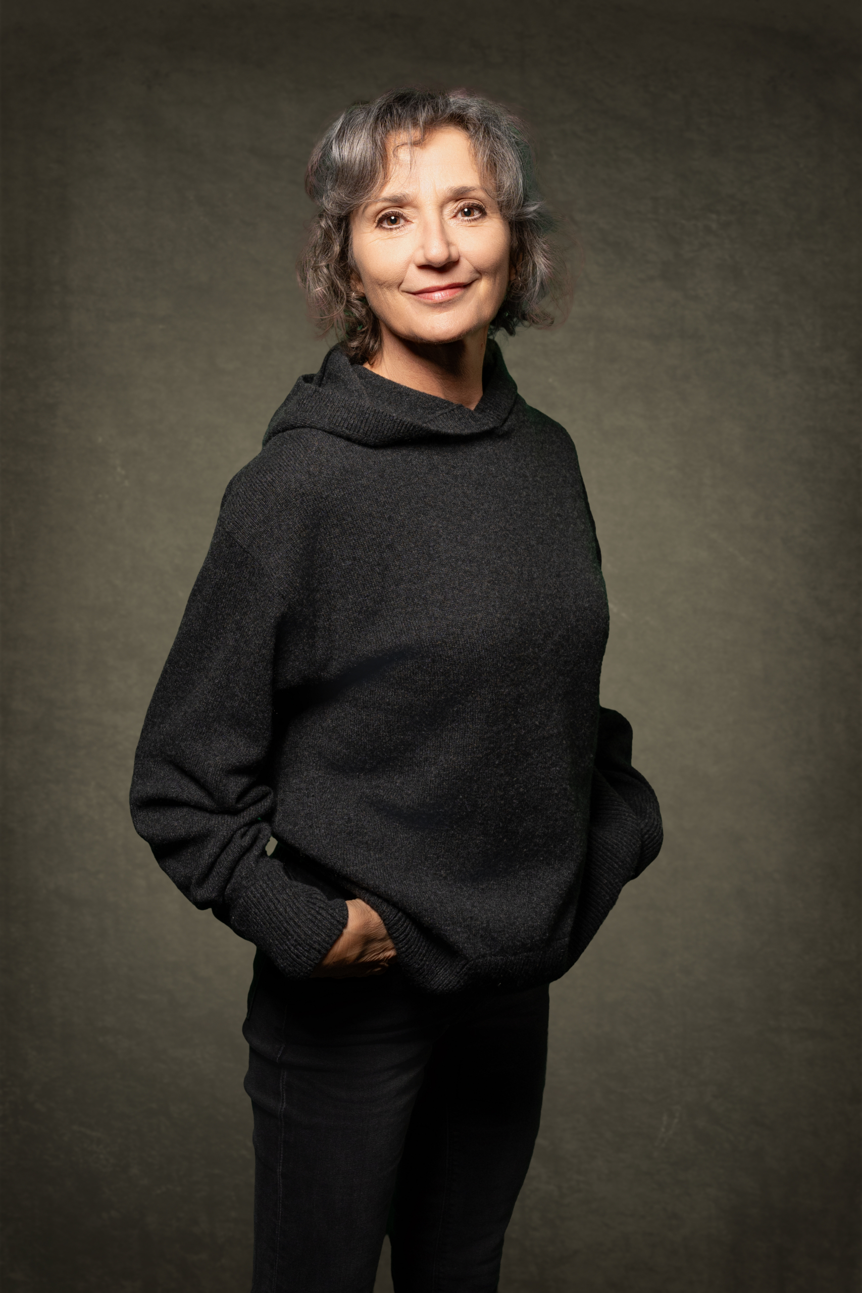 Frau mit grauen Haaren trägt einen schwarzen Hoodie, steht lässig mit Händen in den Taschen.