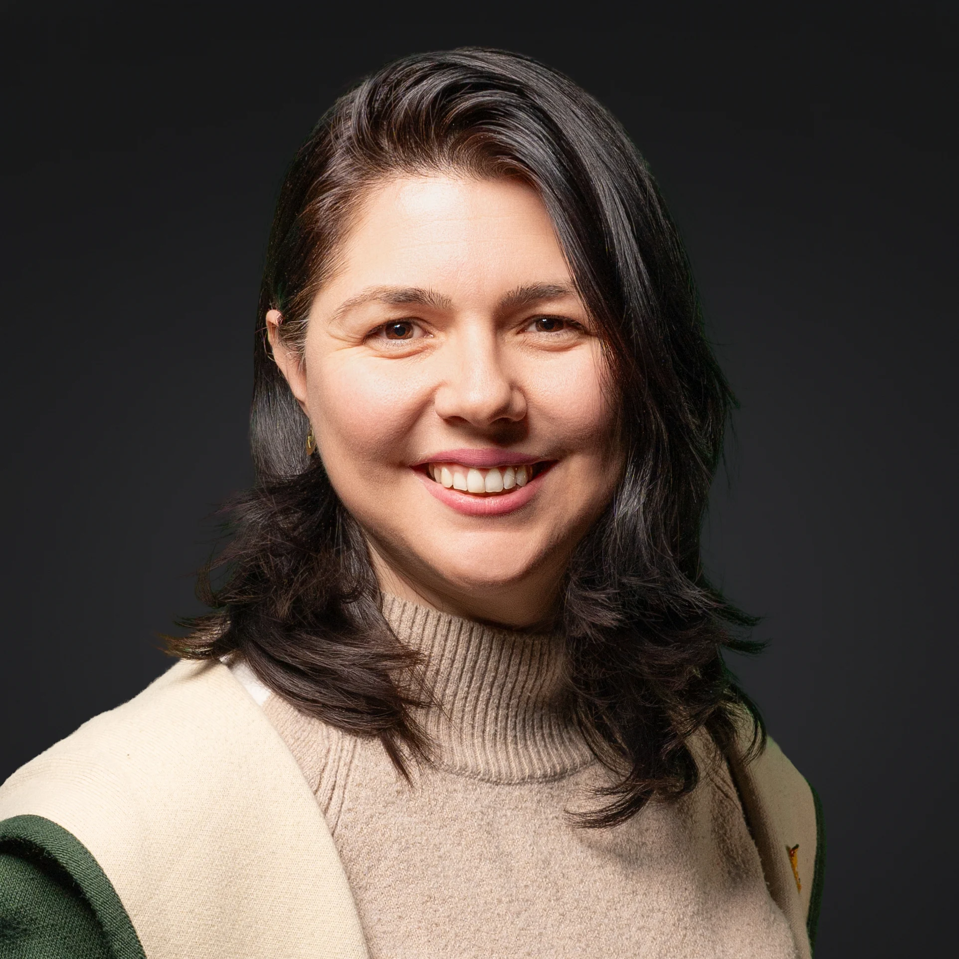 Person mit schulterlangem, dunkelbraunem Haar, trägt einen hellbeigen Rollkragenpullover und eine beige Jacke mit grünen Ärmeln vor dunklem Hintergrund.
