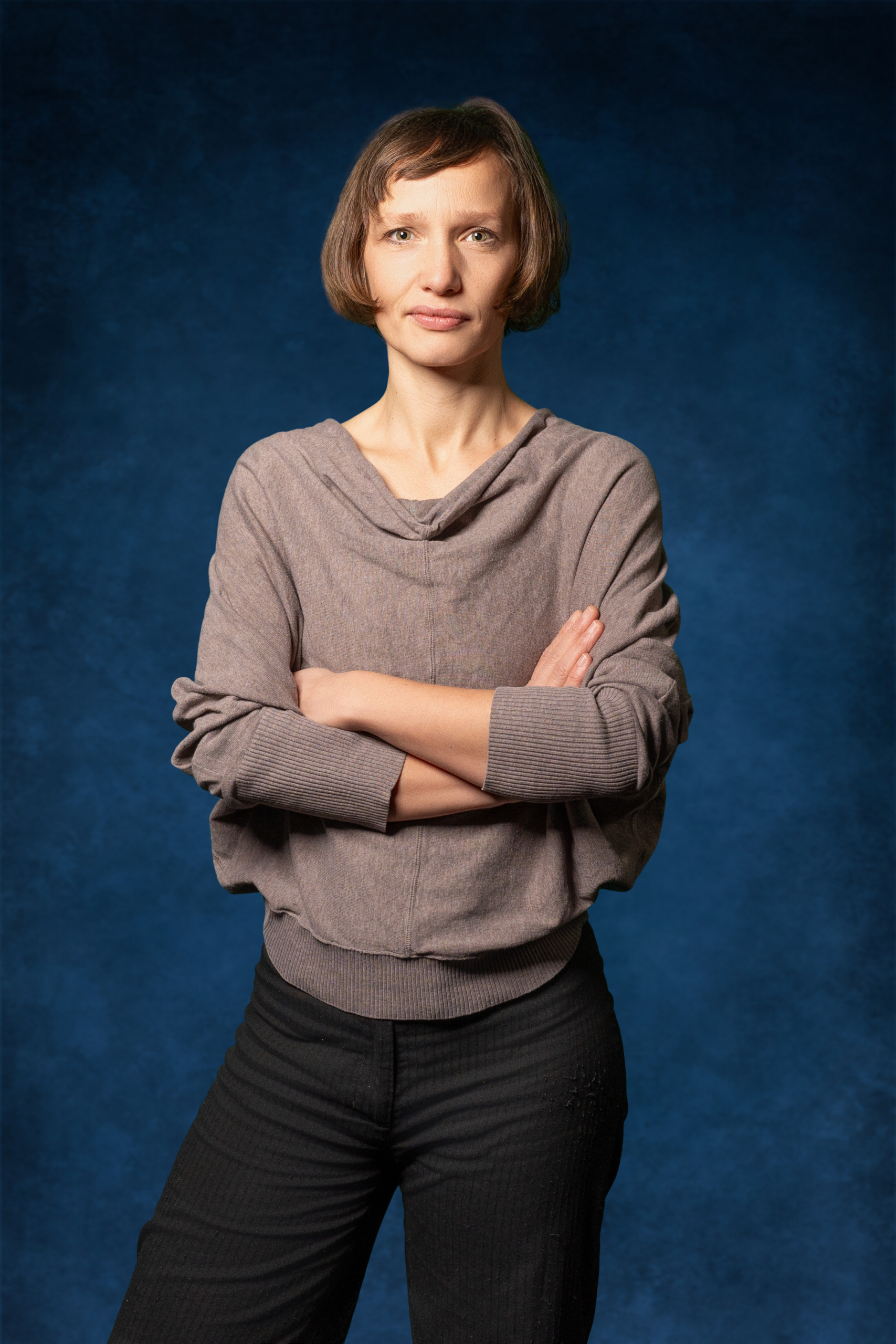Dame mit kurzen Haaren in grauem Pullover und schwarzer Hose posiert vor blauem Hintergrund.