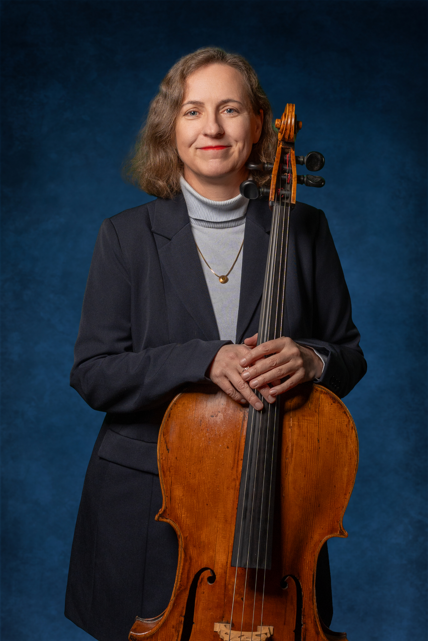 Portrait einer Cellistin mit einem Cello, die in einem Blazer vor einem blauen Hintergrund steht.