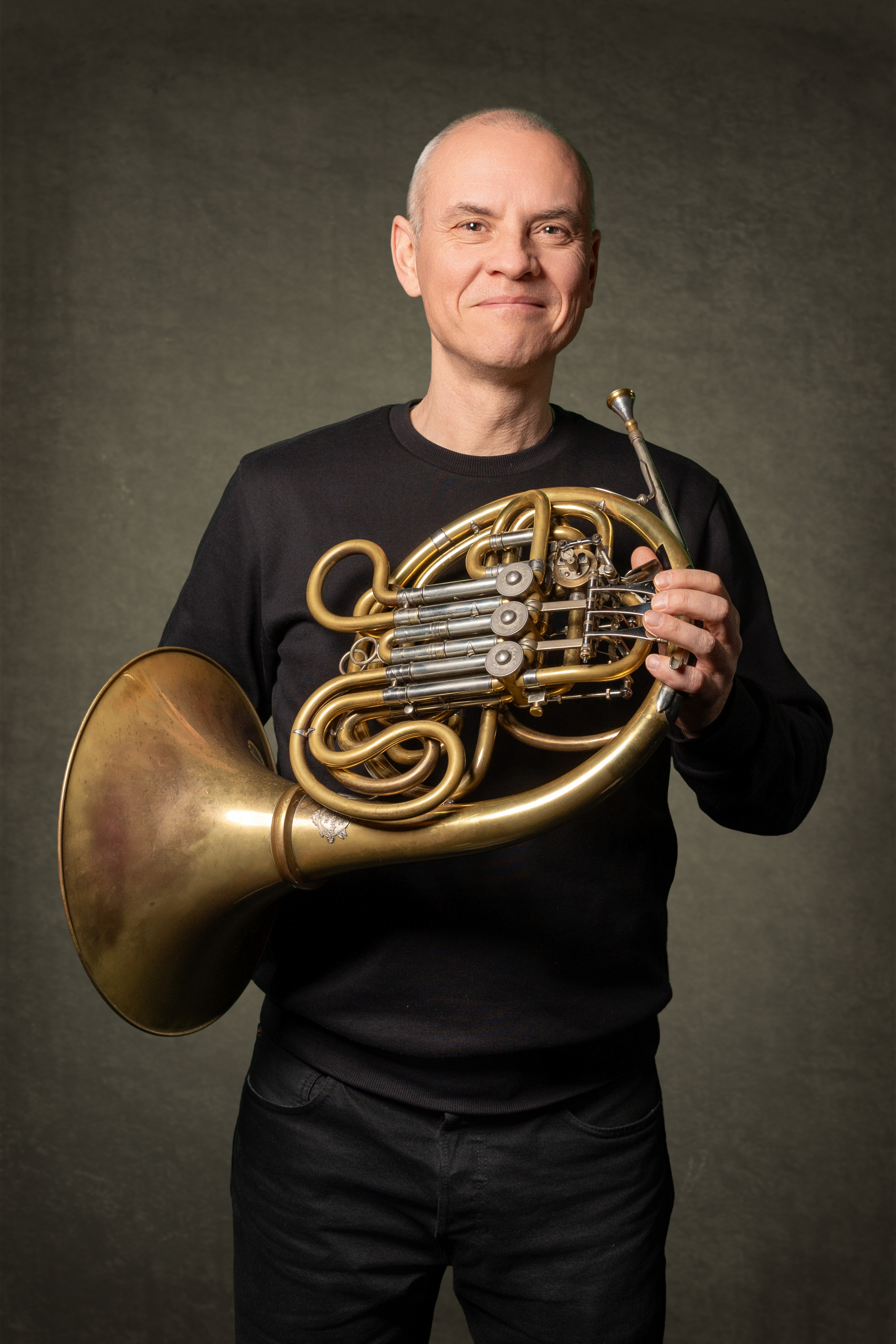 Mann mit Horn in der Hand, lächelt freundlich, steht vor einem grauen Hintergrund.